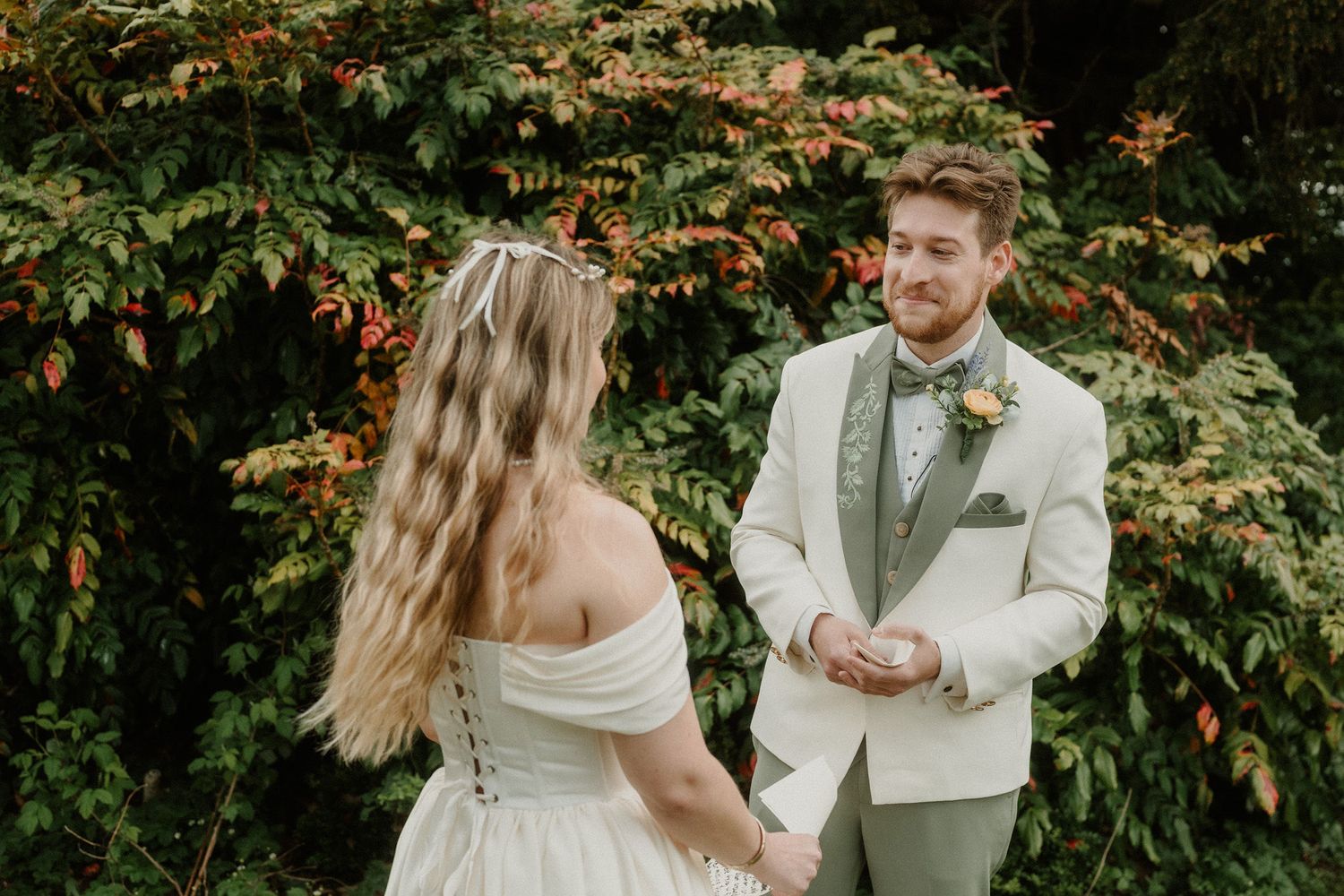 Groom in sage green suit smiles at bride in off-shoulder wedding dress during outdoor ceremony with lush foliage backdrop.