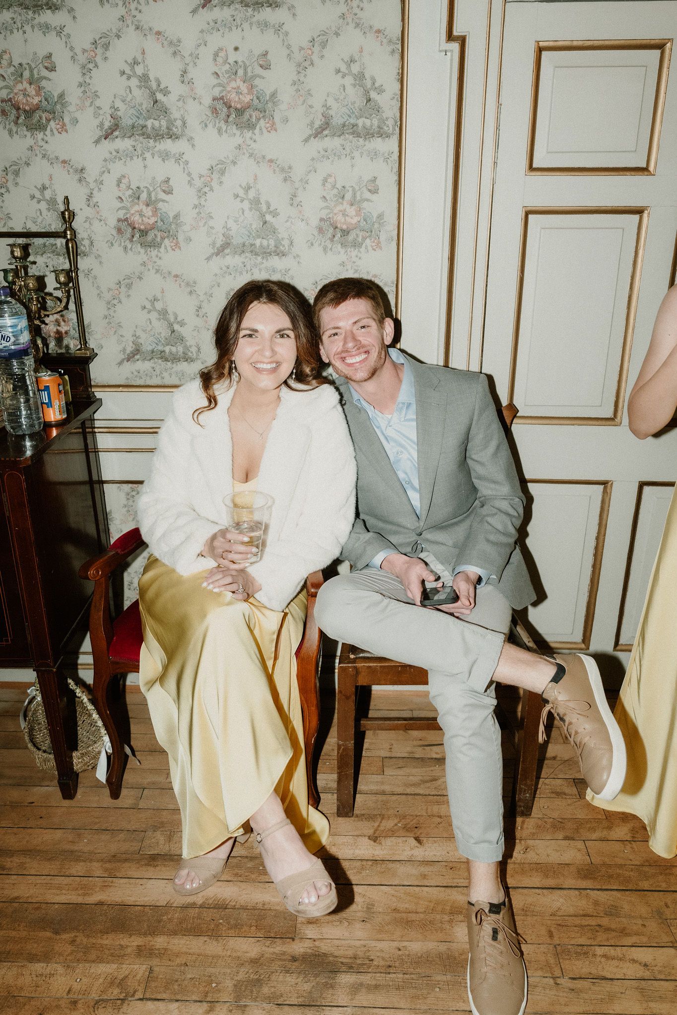 A smiling couple in formal attire sitting together in an elegant room with floral wallpaper and gold trim.