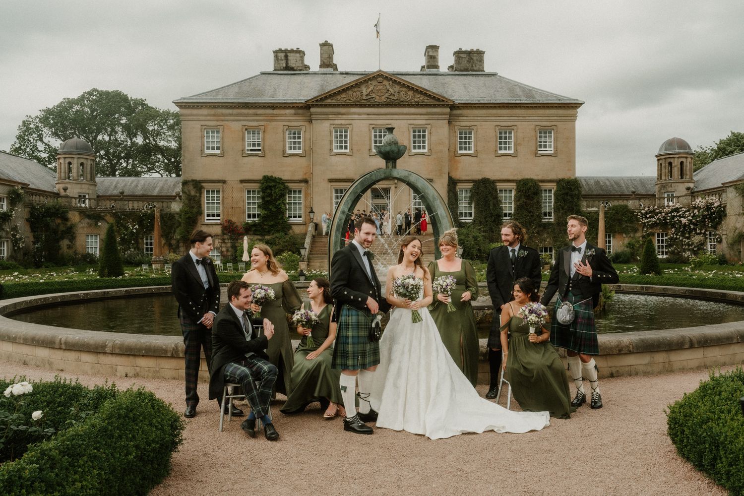 Wedding party poses in front of a grand Scottish manor, groom in tartan kilt, bride in white gown, bridal party in green.