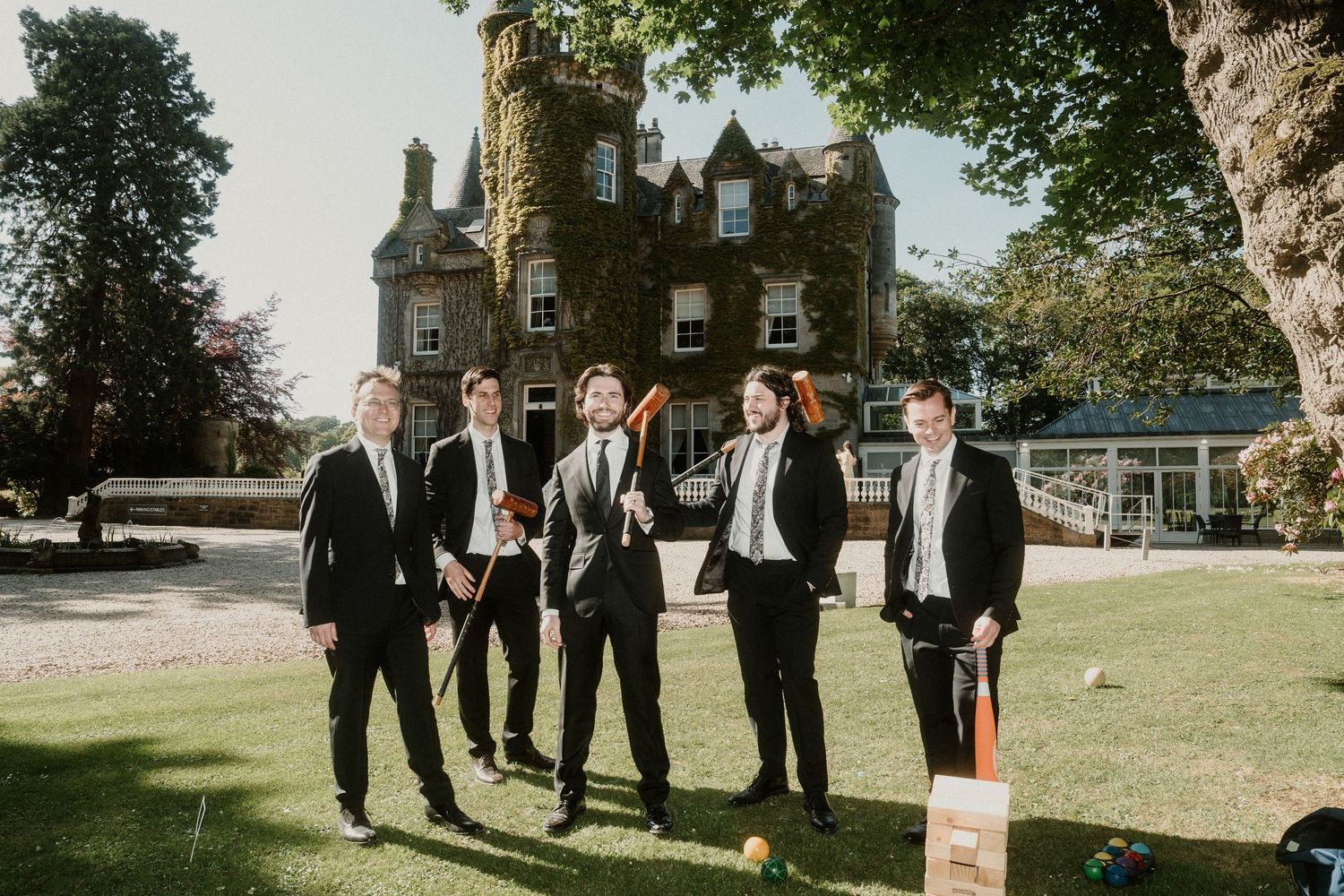 Wedding party members in formal attire engage in outdoor activities on mansion grounds.