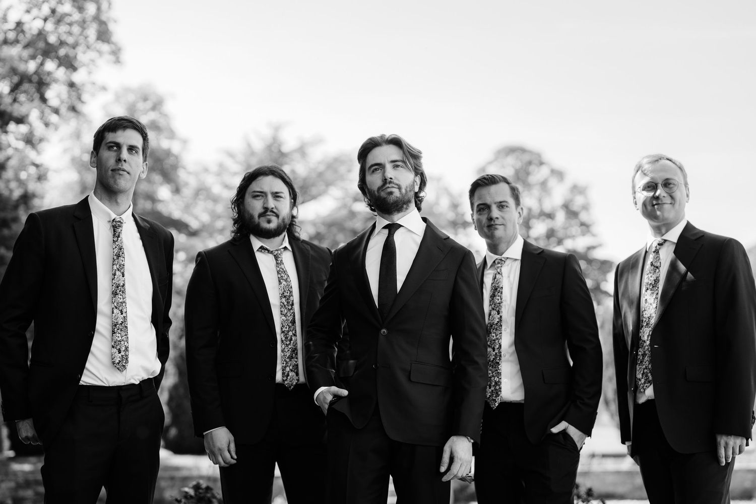 Group in formal black suits standing together outdoors in black and white photography.