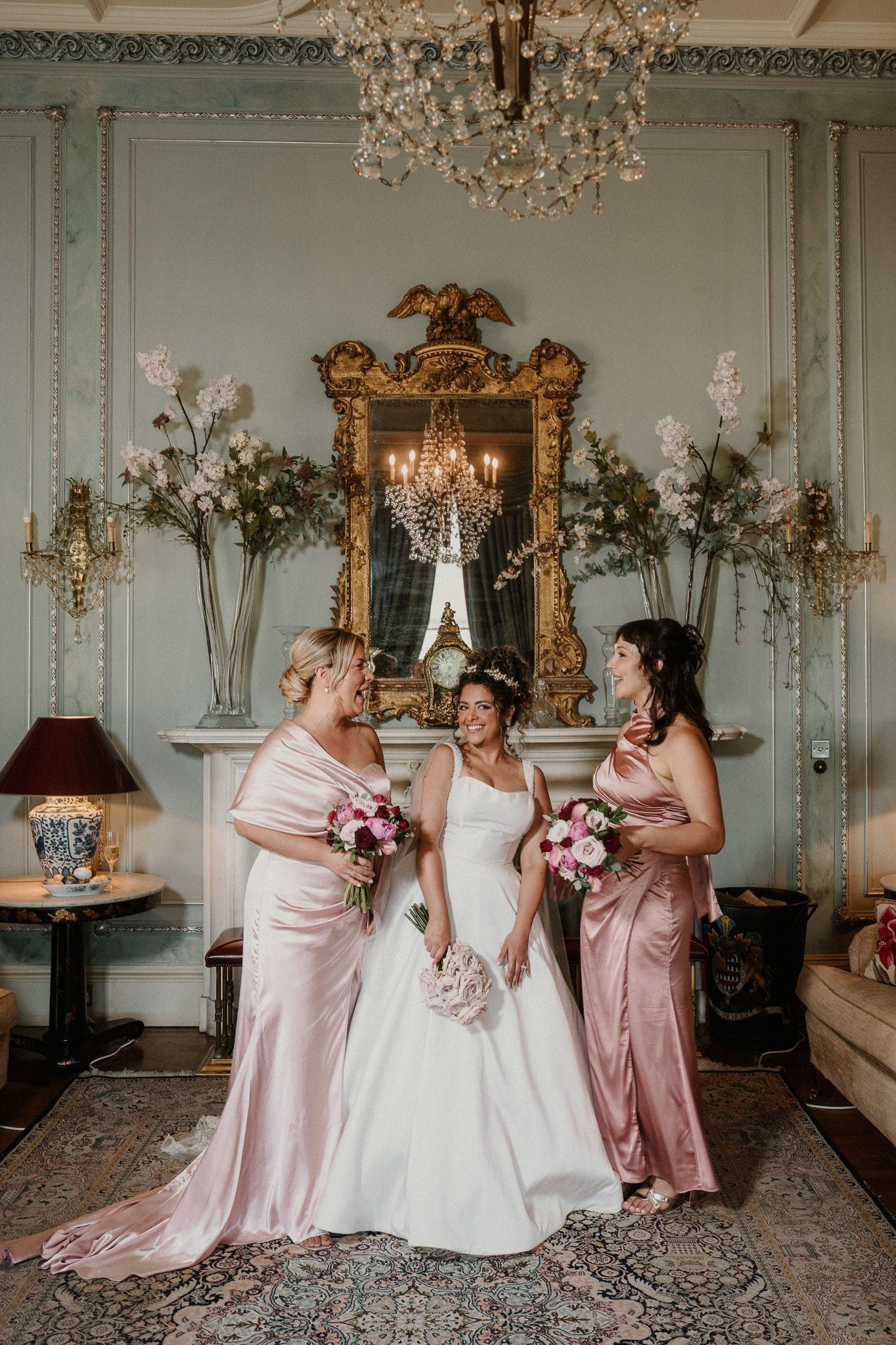 Wedding group shares candid moment in ornate room with gold mirrors and chandeliers.