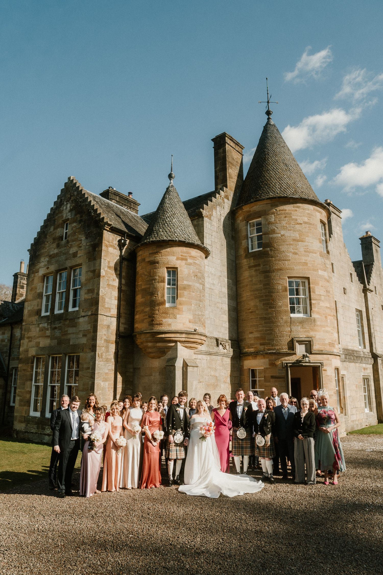 Wedding party poses for group photo outside grand Scottish castle with turrets and stone architecture.