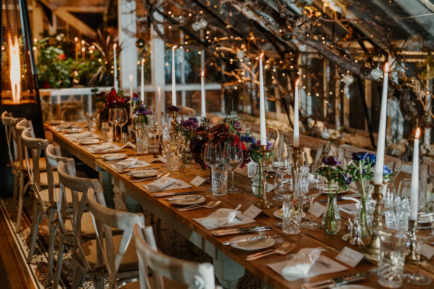 Long wooden table set for a formal dinner with white chairs and tall candles in an indoor garden setting.