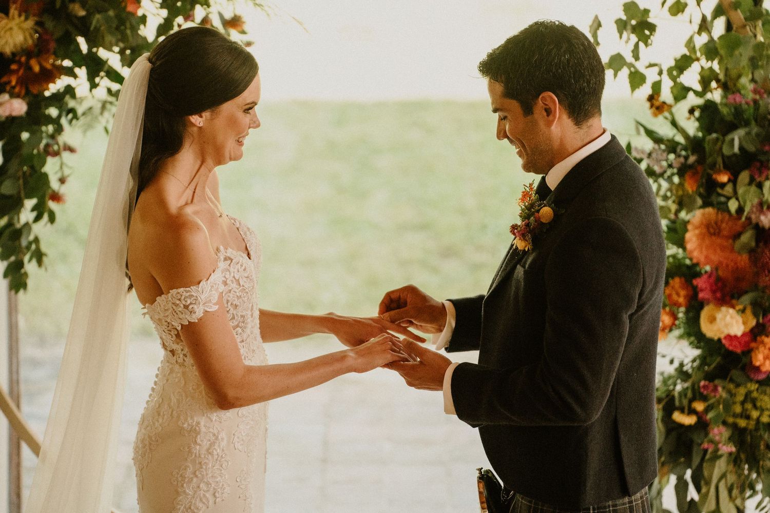 Wedding ceremony exchange under floral arch with warm natural lighting streaming through windows.