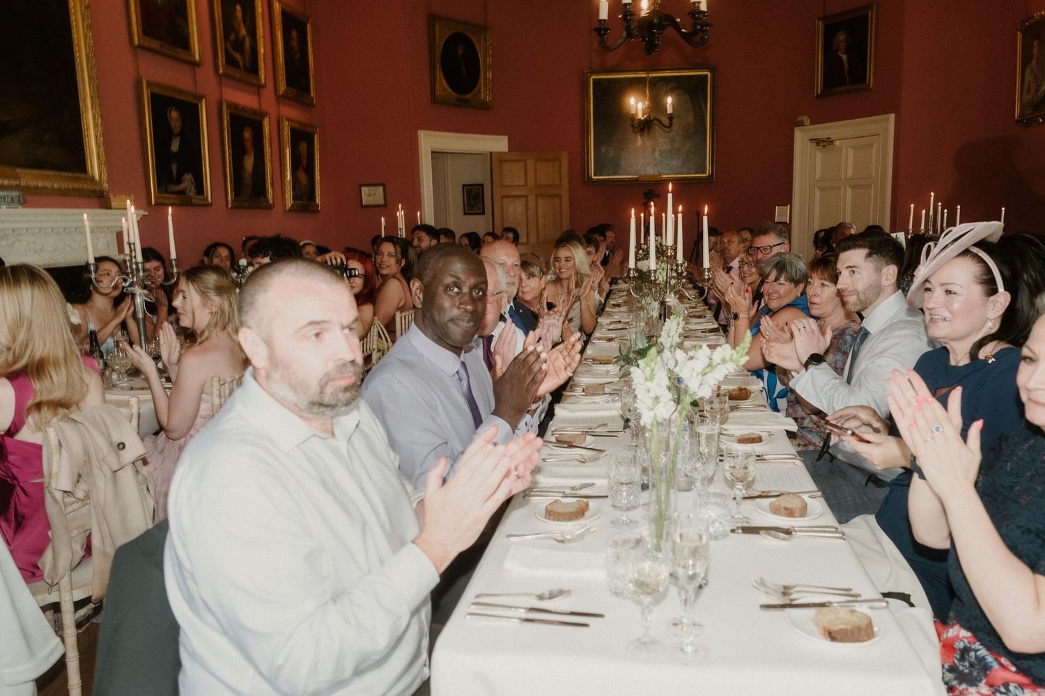 Guests applauding at an elegant formal dinner in a grand red-walled dining room with candlelit tables.
