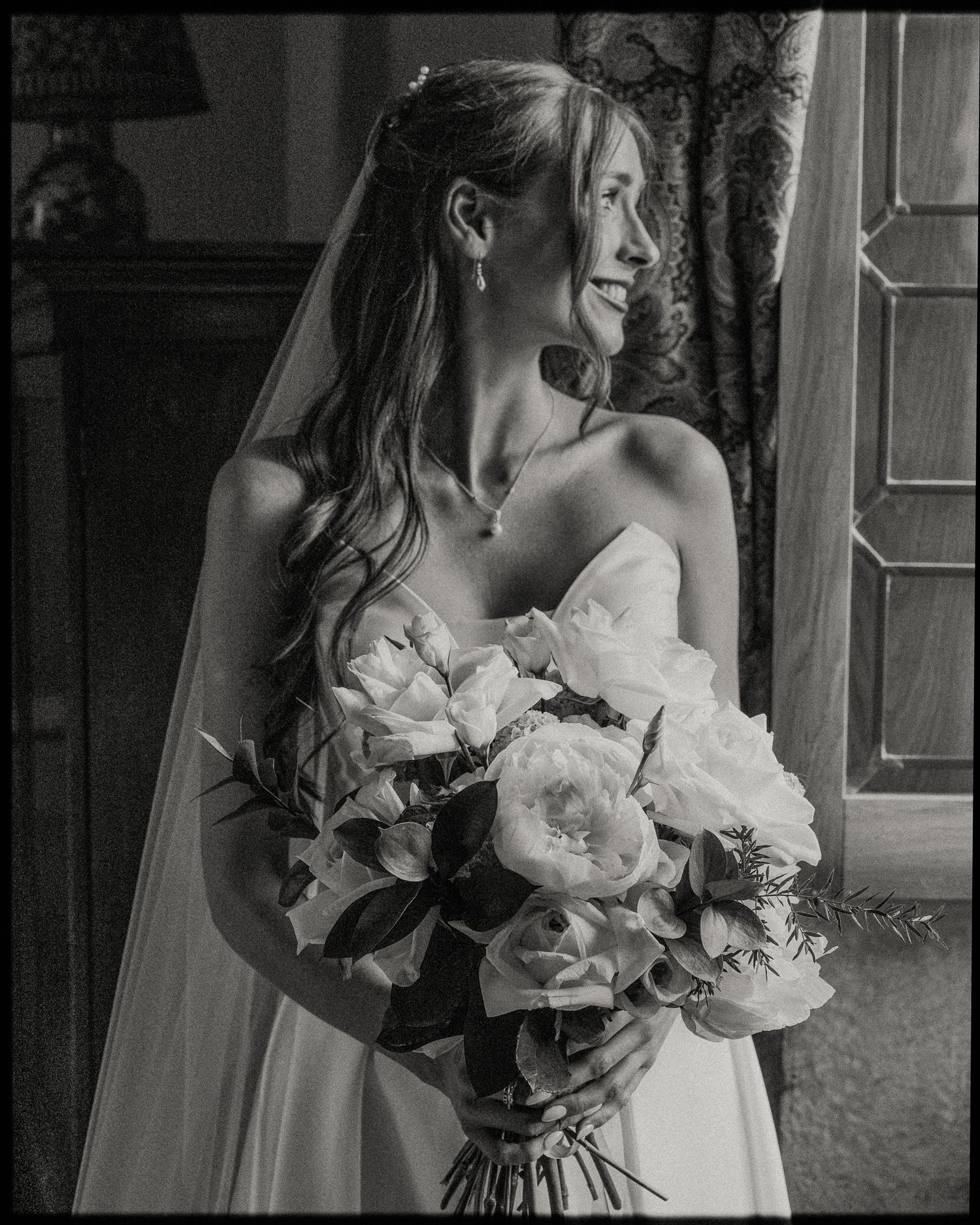 Elegant bride in strapless gown holding white rose bouquet, smiling in black and white portrait photo.