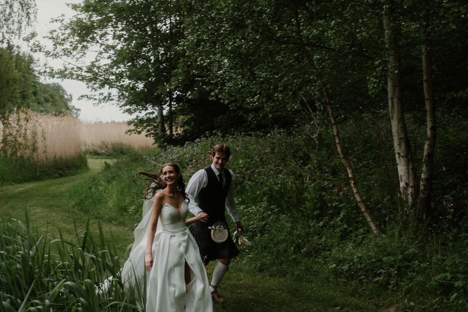A bride in white gown and groom in Scottish kilt walk together along a lush green woodland path.