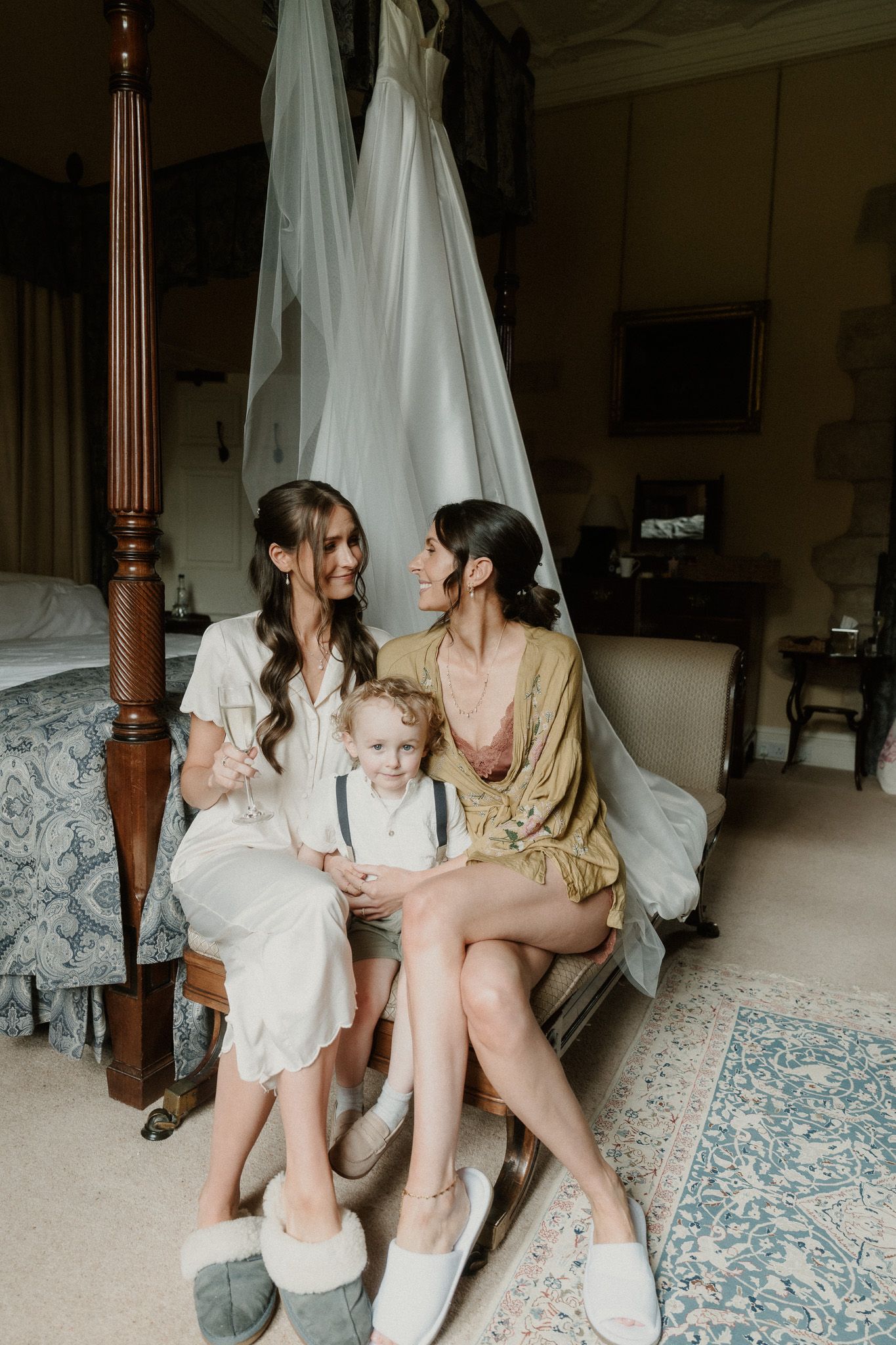 Two bridesmaids in robes sit on a bed with a young boy, smiling, as a wedding dress hangs behind them.