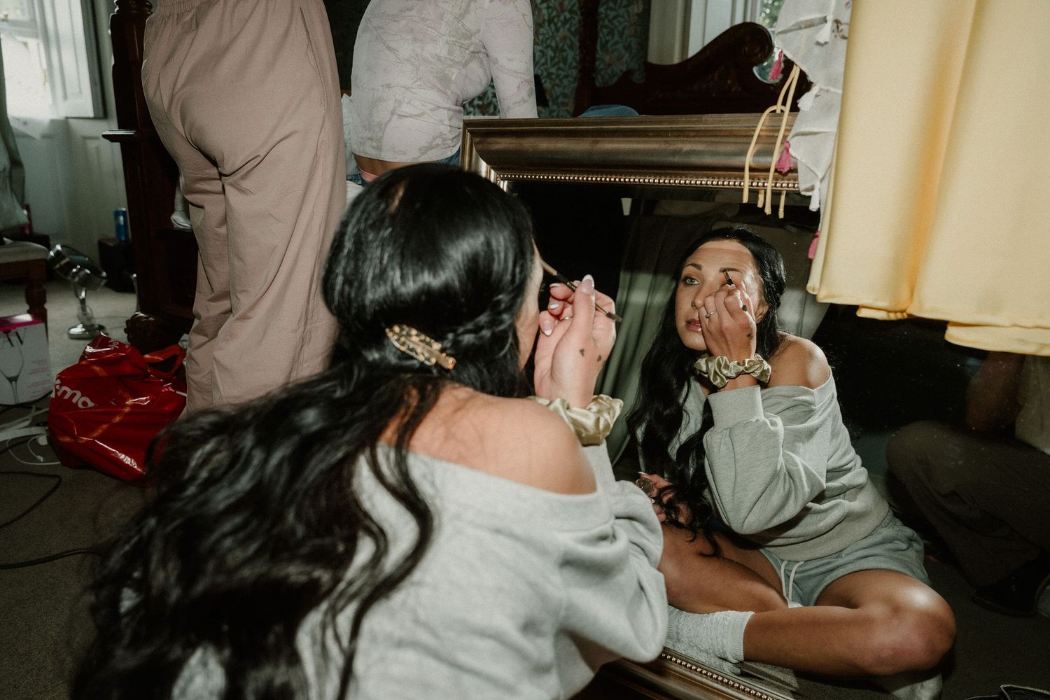 A makeup artist applies makeup to a seated person in a casual indoor setting with warm lighting.