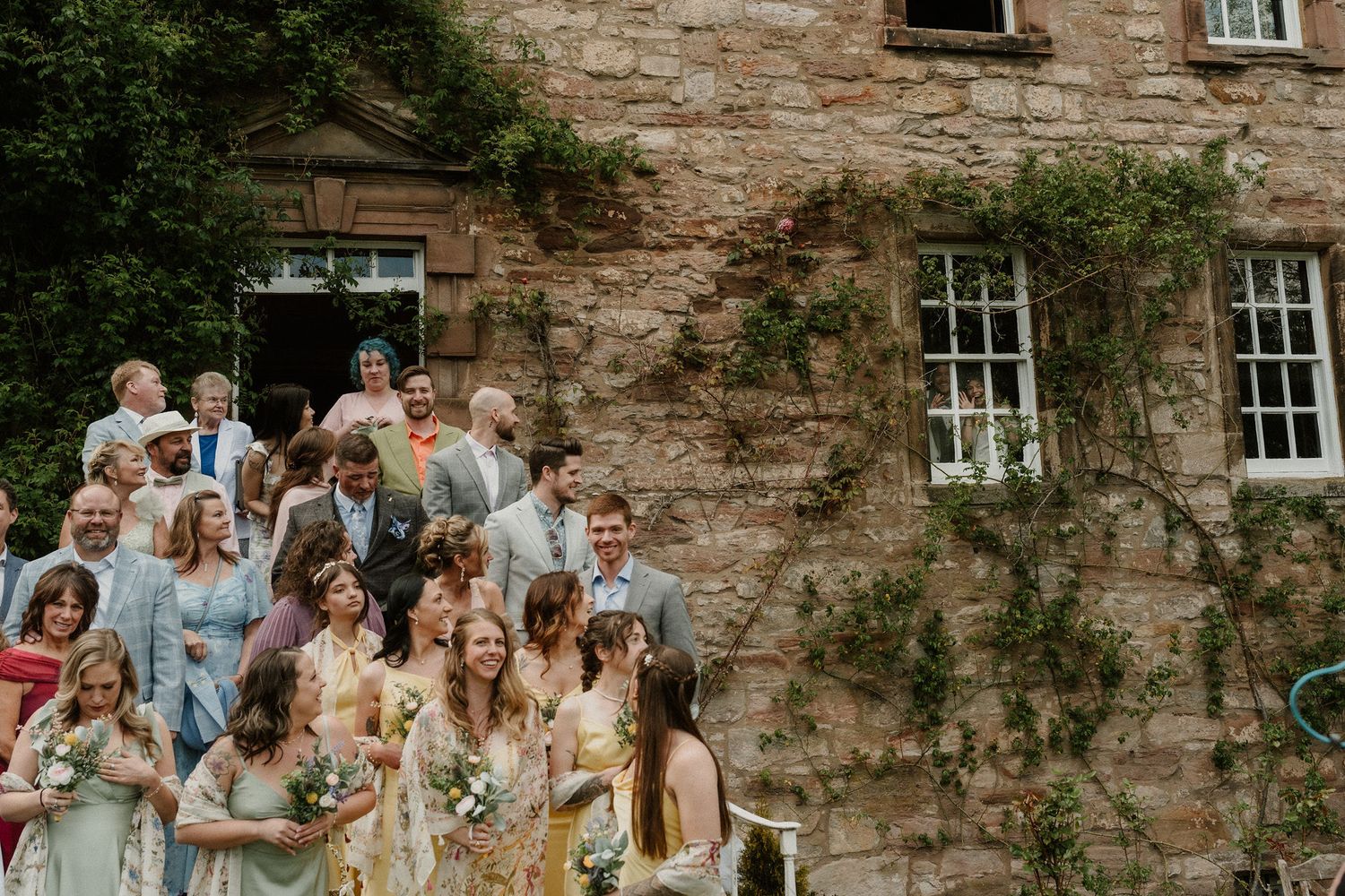 Wedding guests gathered outside a rustic stone manor covered in ivy, celebrating in colorful summer attire.