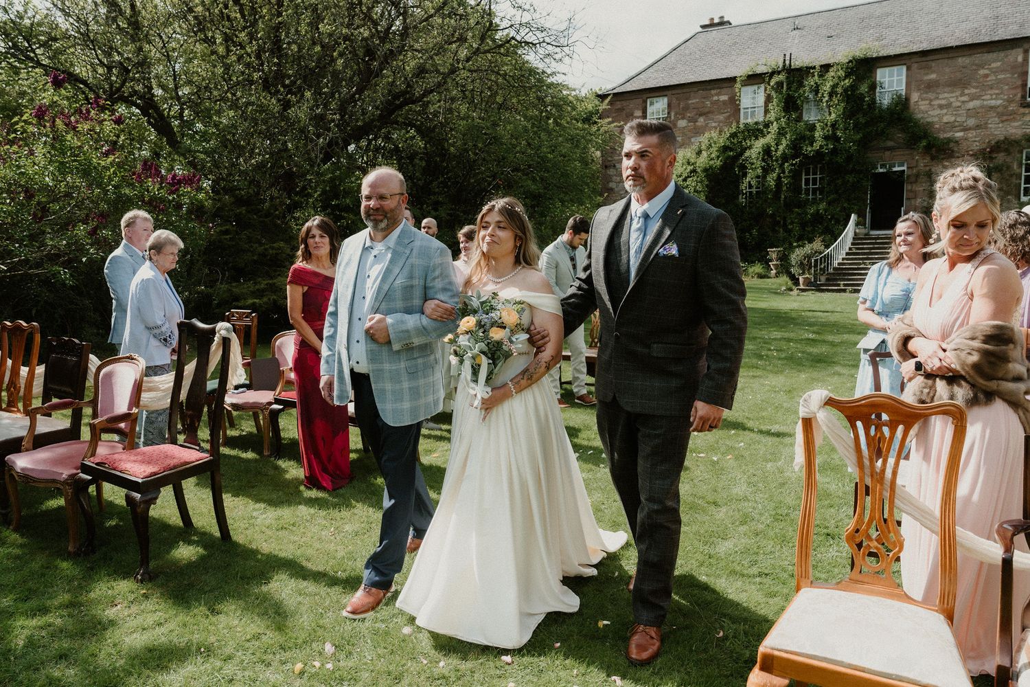 Bride walks down outdoor aisle flanked by two men, guests seated on wooden chairs, stone building backdrop.