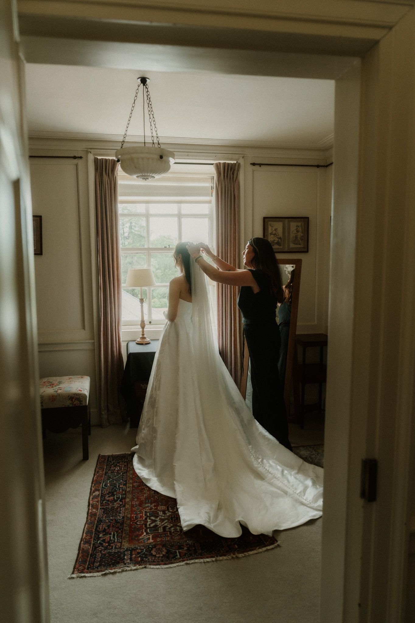Bride in elegant white gown with long veil being adjusted by bridesmaid near window in classic room.