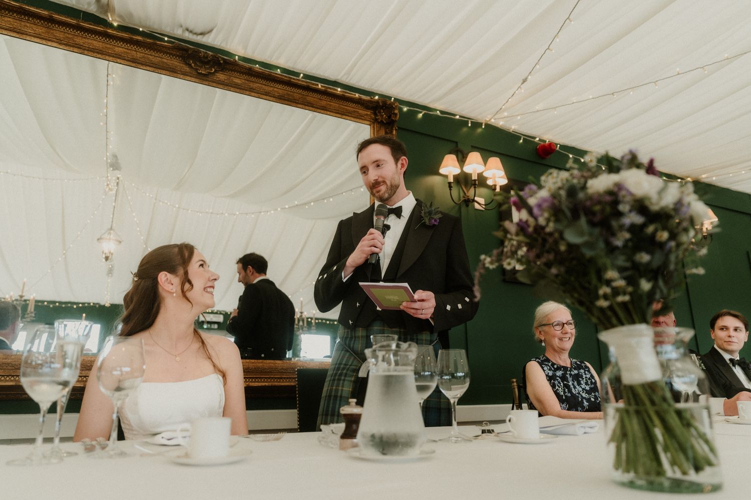 Groom gives a heartfelt speech at an elegant wedding reception in a marquee tent with floral decorations.