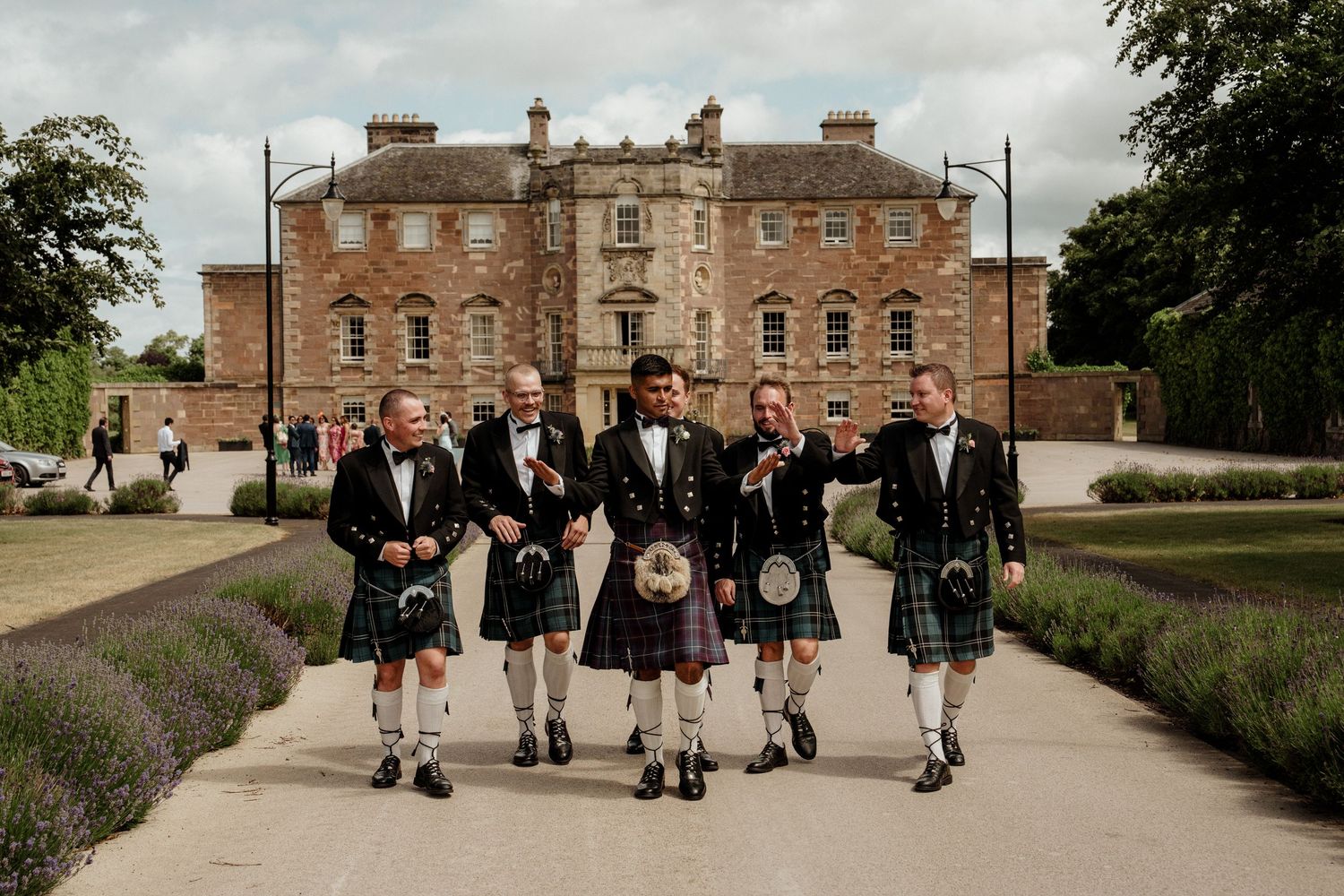 A group in formal attire and kilts walking along a path outside a historic brick building.