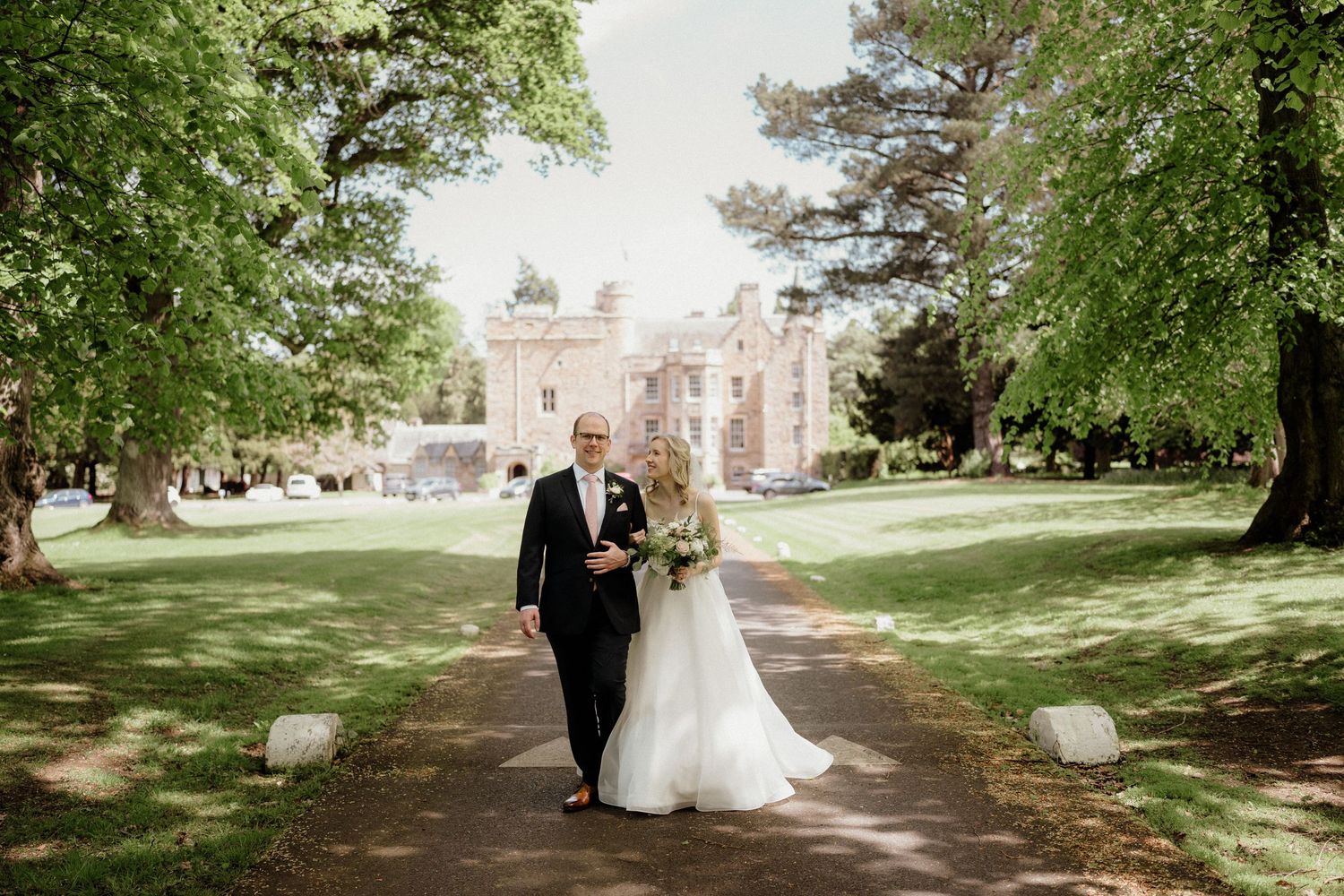 A romantic wedding couple strolls along a tree-lined path on a sunny day at an elegant estate.