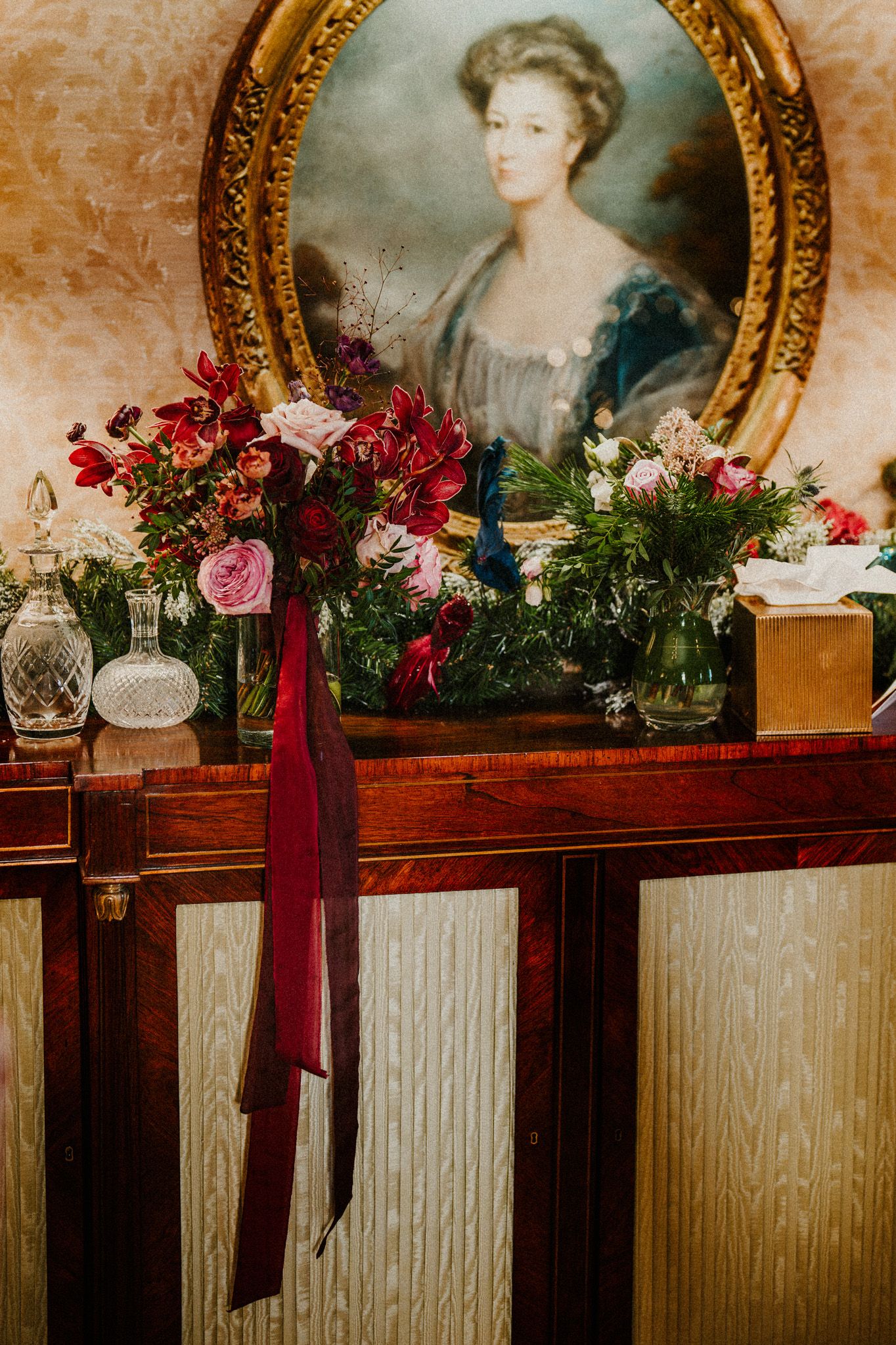 Vintage-style mantel decorated with red and pink floral arrangements beneath antique gold-framed portraits.