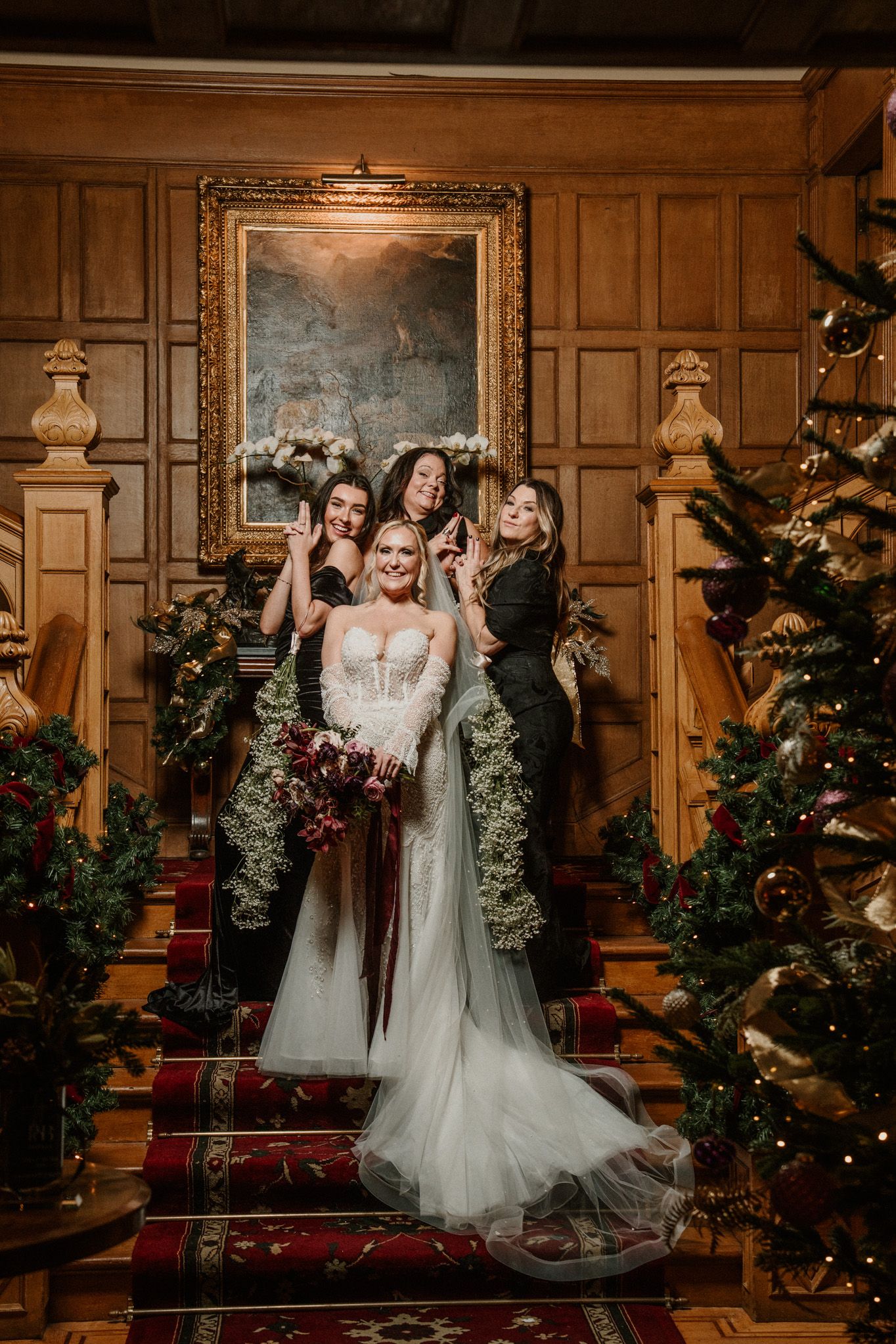 Wedding party poses on elegant wood-paneled staircase with decorated Christmas trees and garland for holiday celebration.