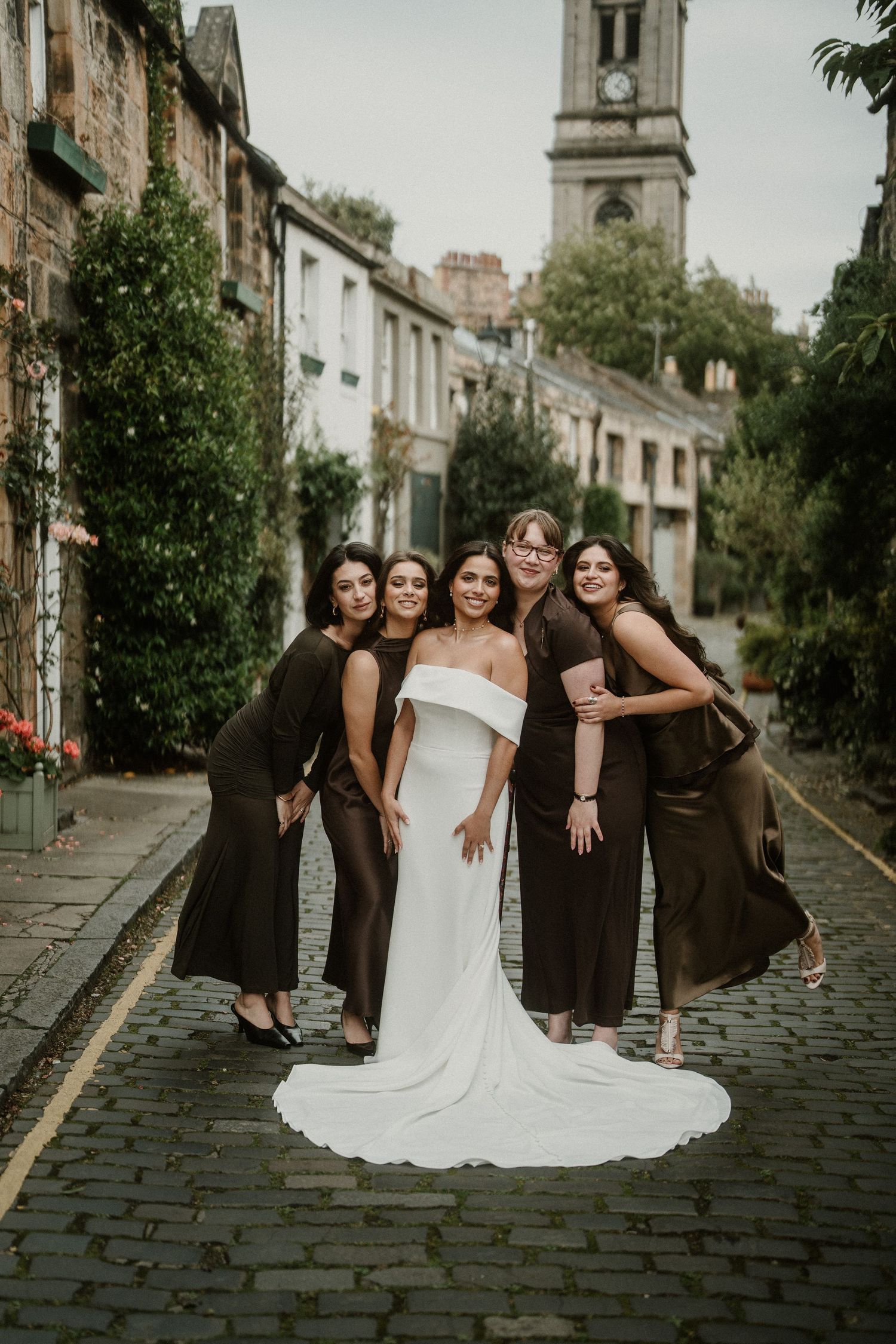 Bridal party in black dresses and white gown pose together on historic cobblestone street.