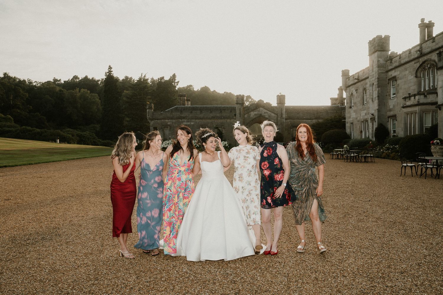 A wedding party poses on the grounds of a grand estate with a bride in white and attendants in colorful dresses.