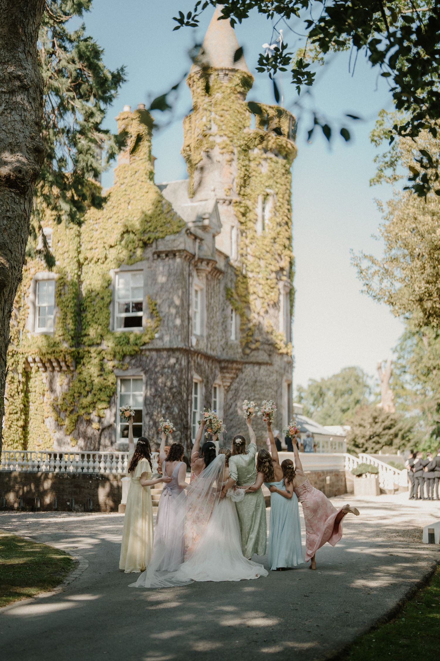Bridesmaids in soft-colored dresses gather outside historic stone mansion on sunny day.