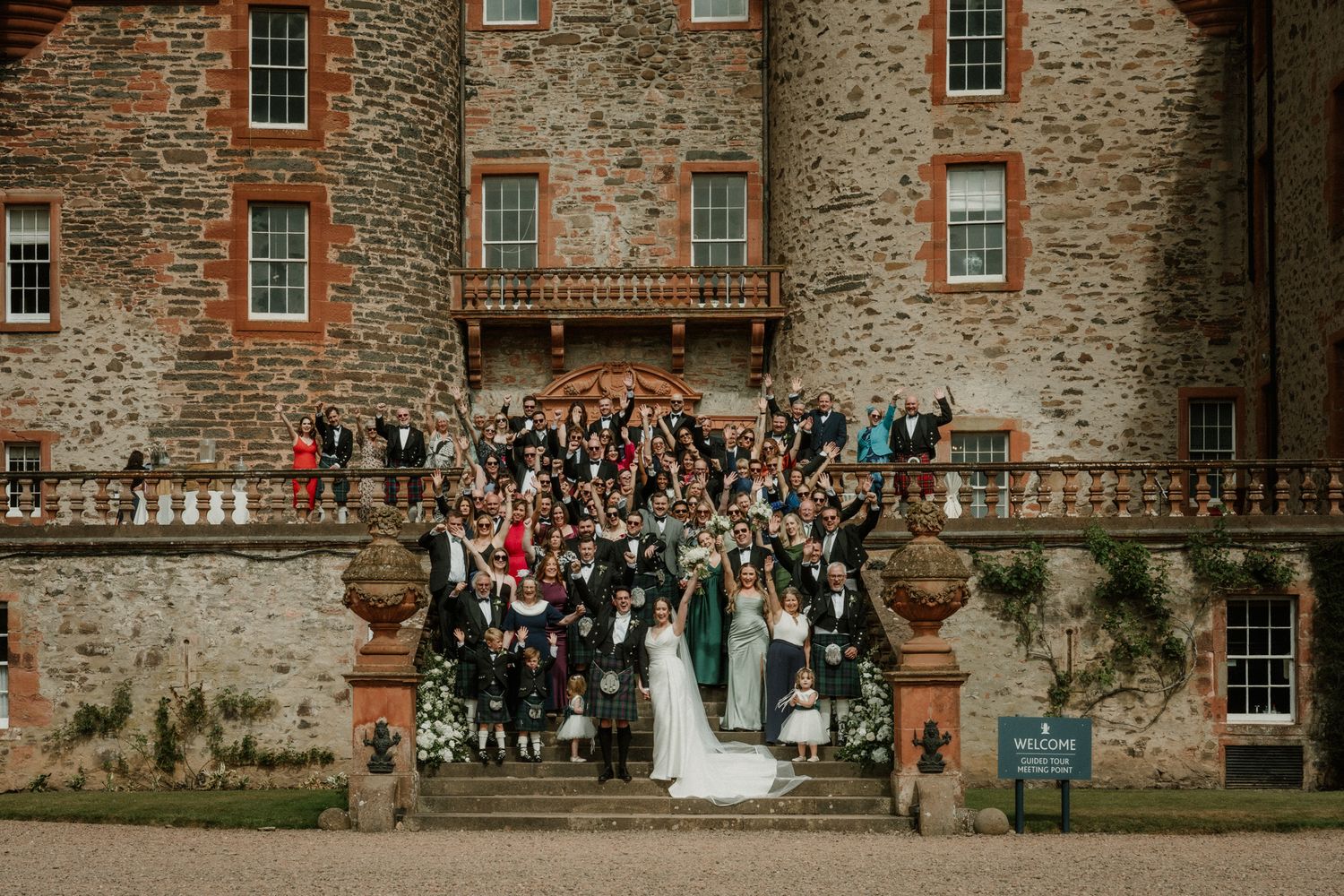 Large wedding party gathered on stone castle steps around a bride in a white gown, with guests in formal attire and kilts.