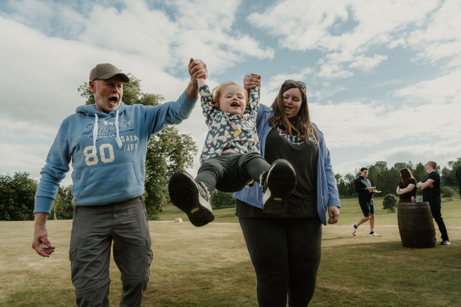Two smiling adults swing a laughing toddler between them while walking across a sunny grassy field with trees and people in the background.