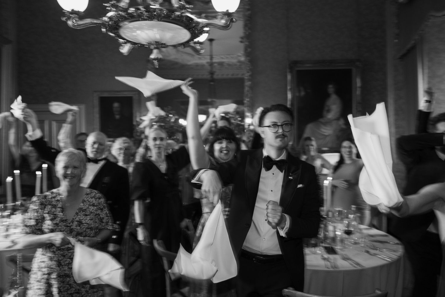 Guests in formal attire cheer and toss paper airplanes at an elegant candlelit banquet in a grand ballroom, black and white photo.