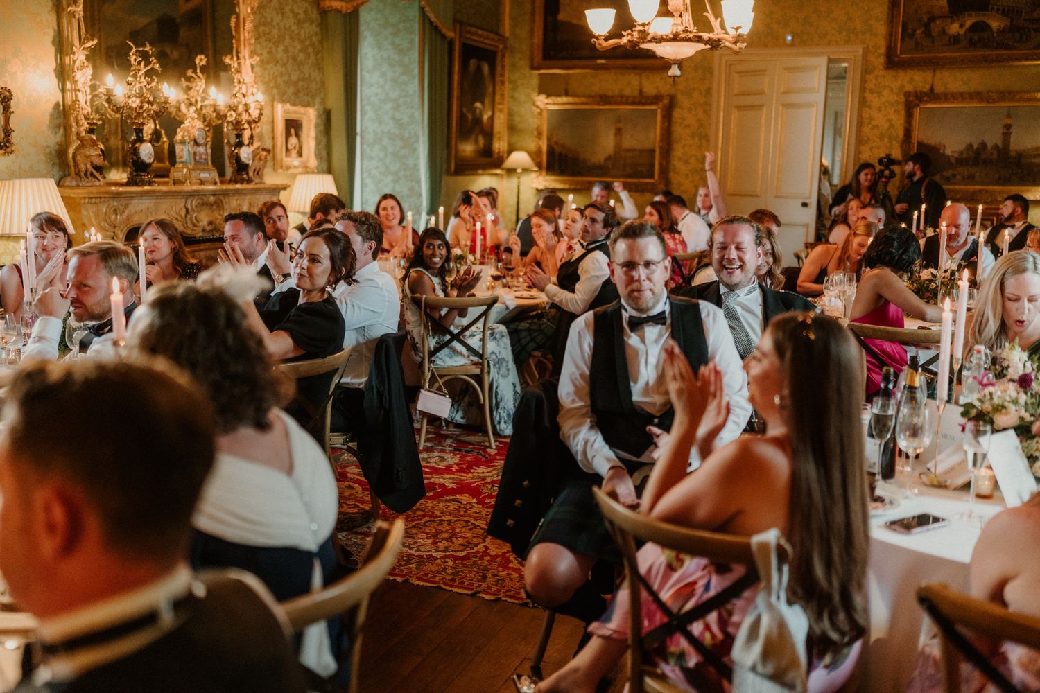 Wedding guests in formal attire laugh and enjoy dinner at candlelit tables in an elegant historic venue with ornate chandeliers.
