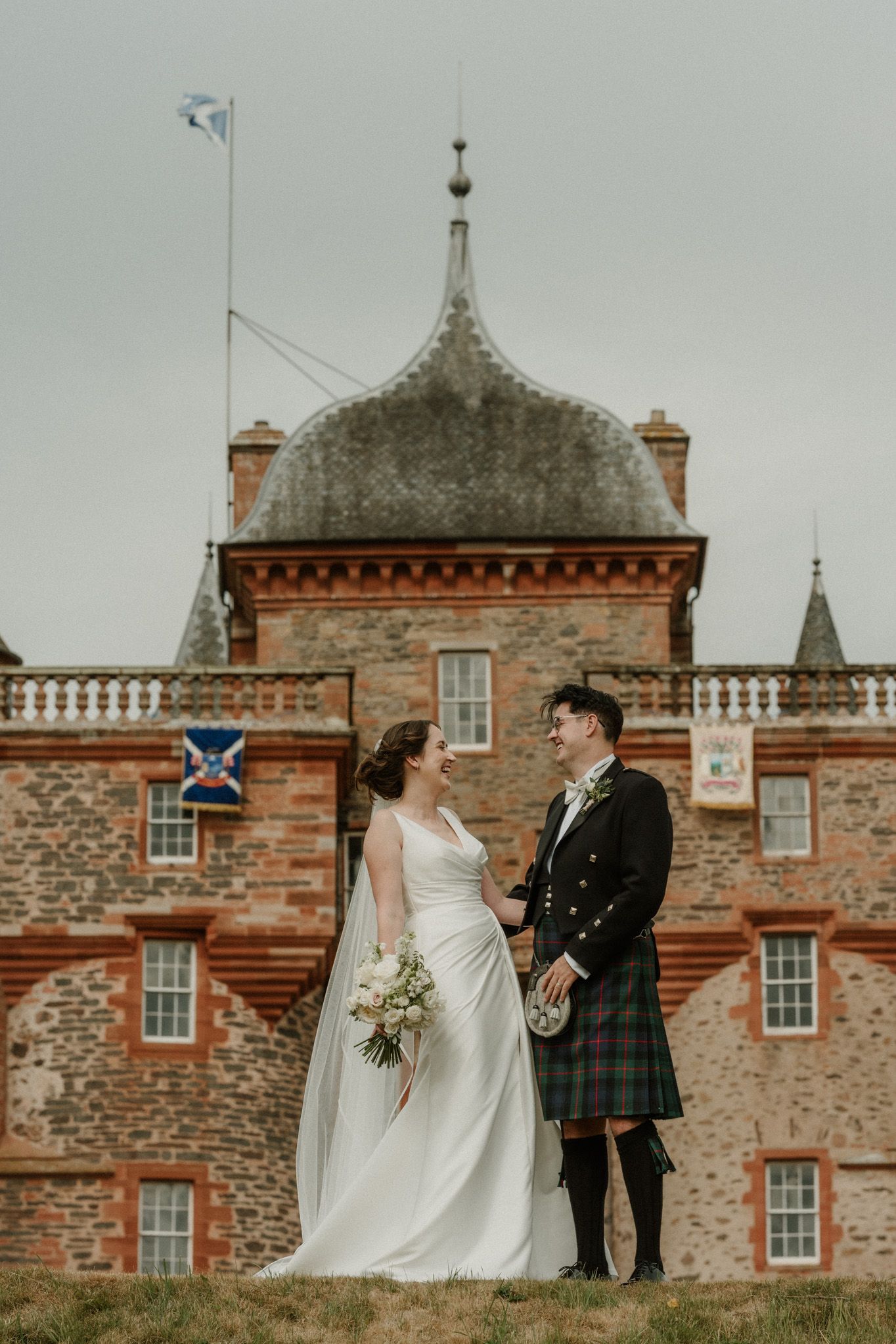 Bride in a white gown and groom in a tartan kilt laugh together in front of a historic red sandstone Scottish castle.