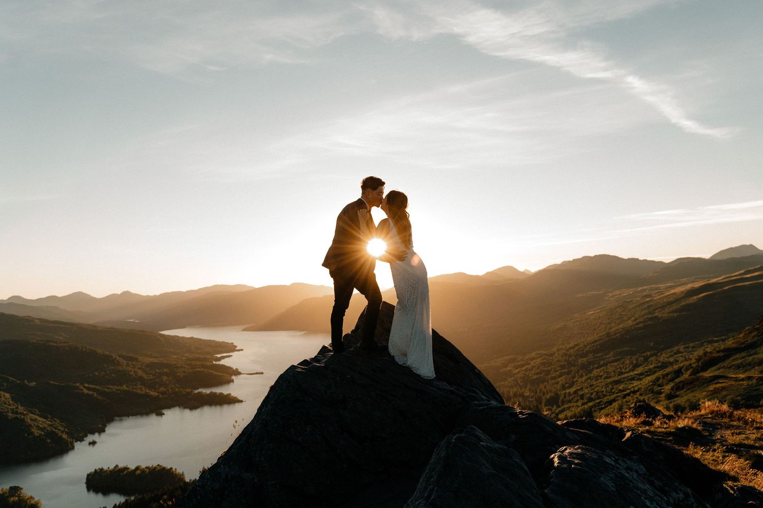 Couple silhouetted against golden sunset on mountain peak, sharing a kiss with scenic loch and hills in background.