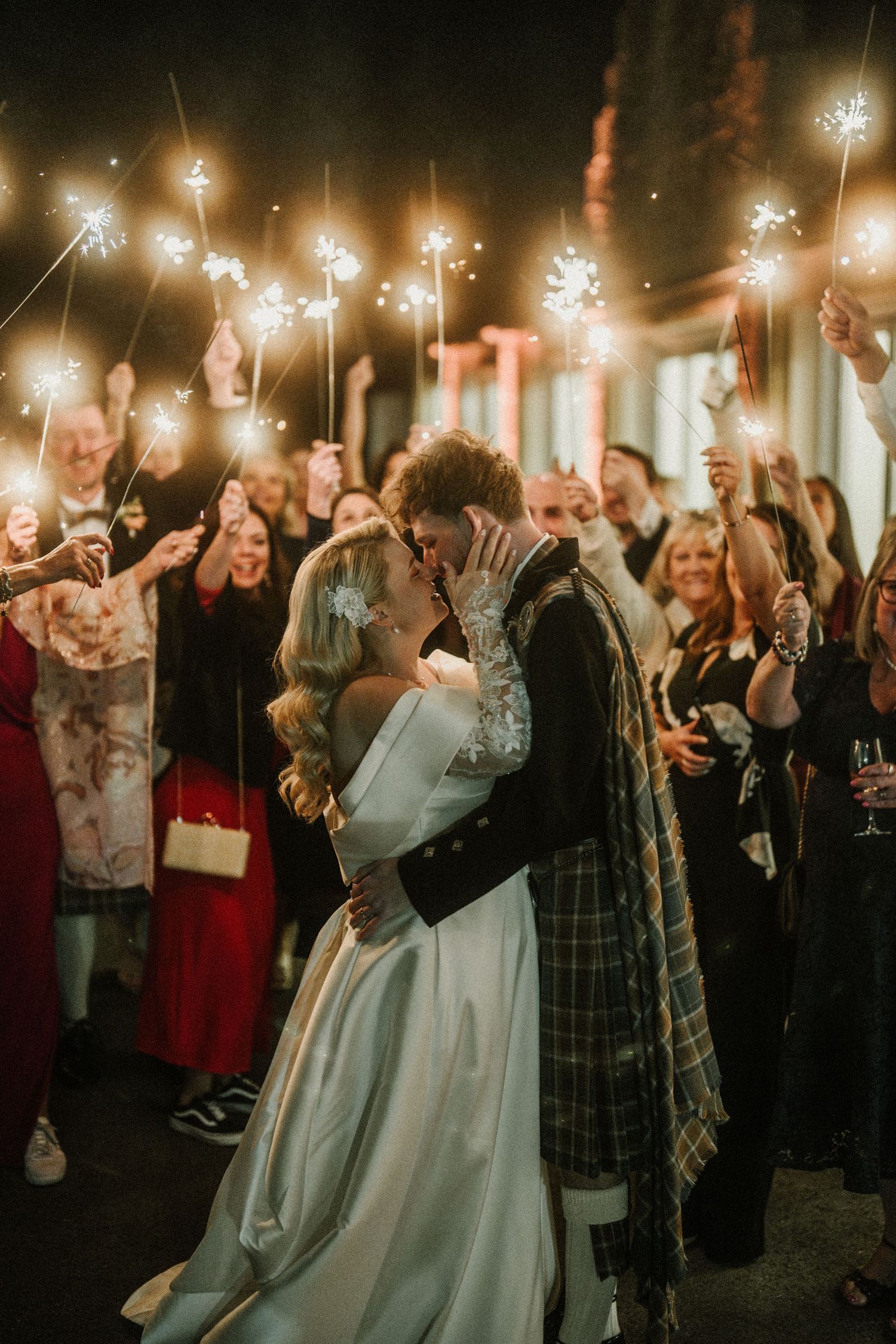 A newlywed couple shares a romantic kiss surrounded by guests holding sparklers at a Scottish wedding celebration.