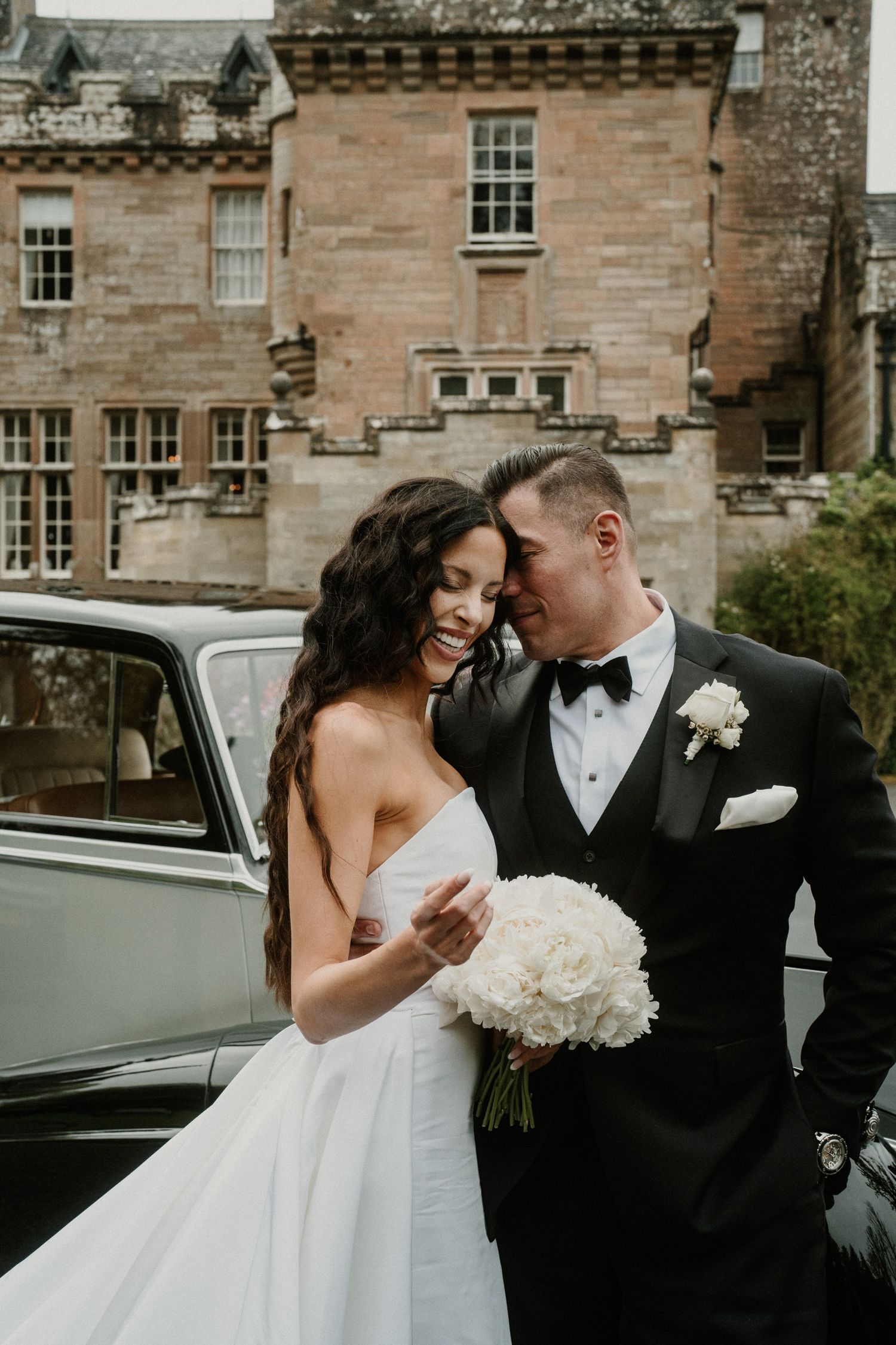 A romantic wedding portrait in front of a historic stone castle with a classic car.