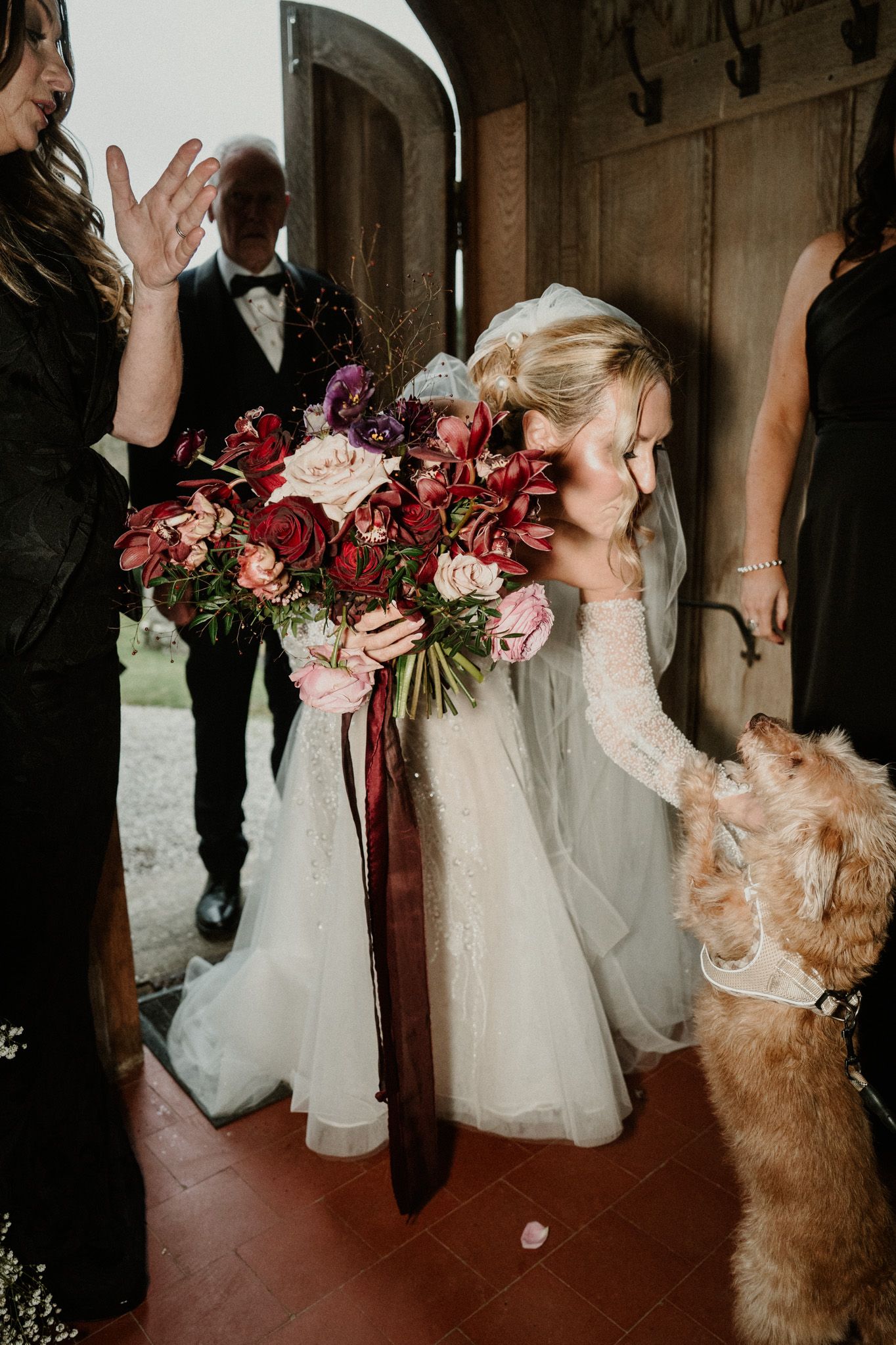 A bride in a white gown bends down to greet an excited dog while holding a burgundy bouquet.