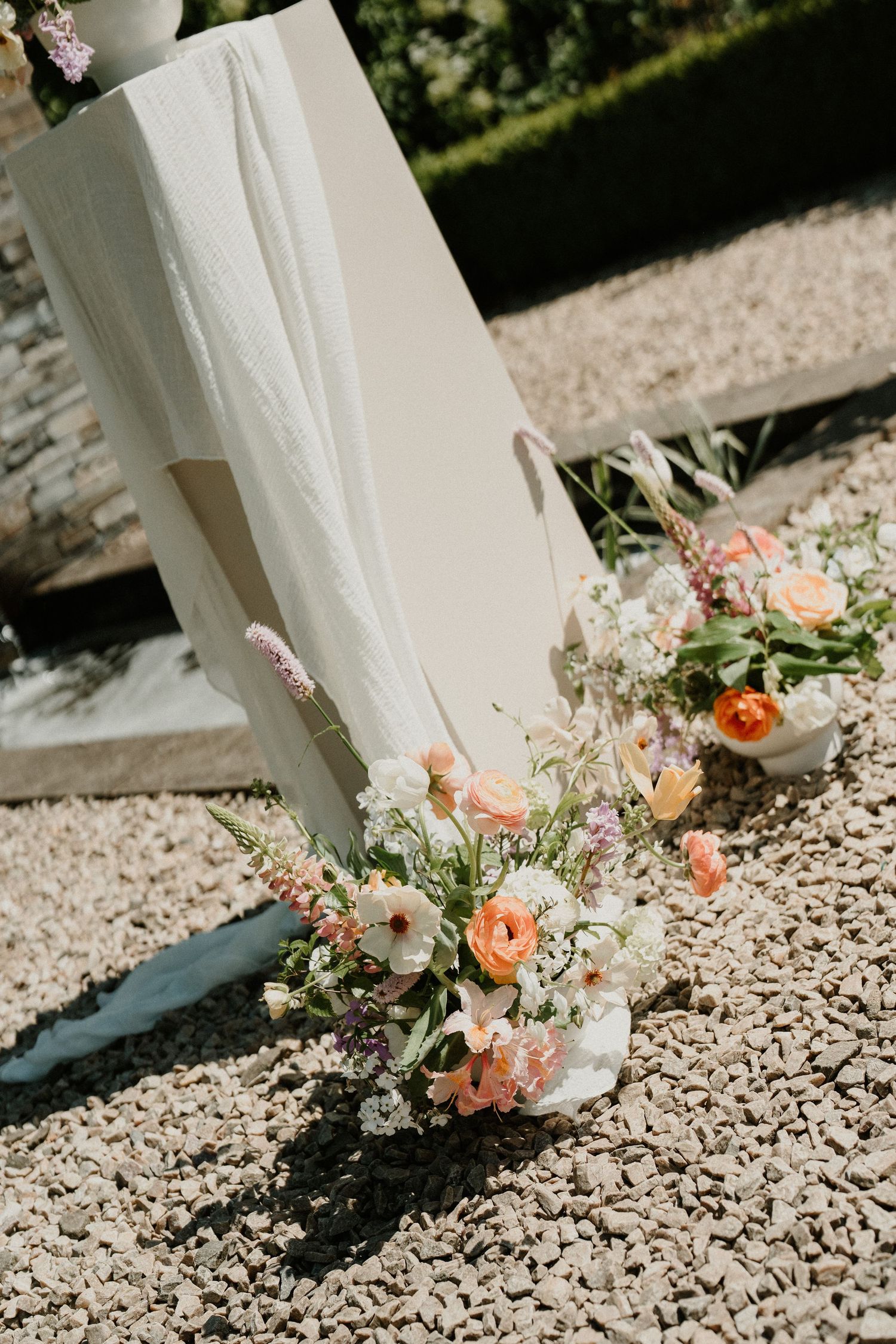 Close-up of romantic peach and white flower arrangements on gravel with flowing white fabric.