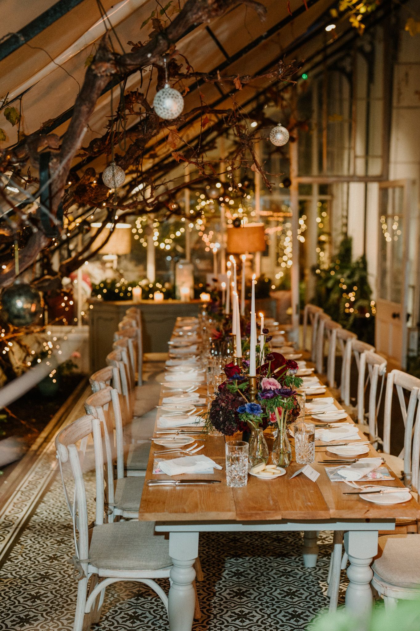 Rustic-elegant dining setup with white chairs and moody floral centerpieces under hanging glass orbs and twinkle lights.