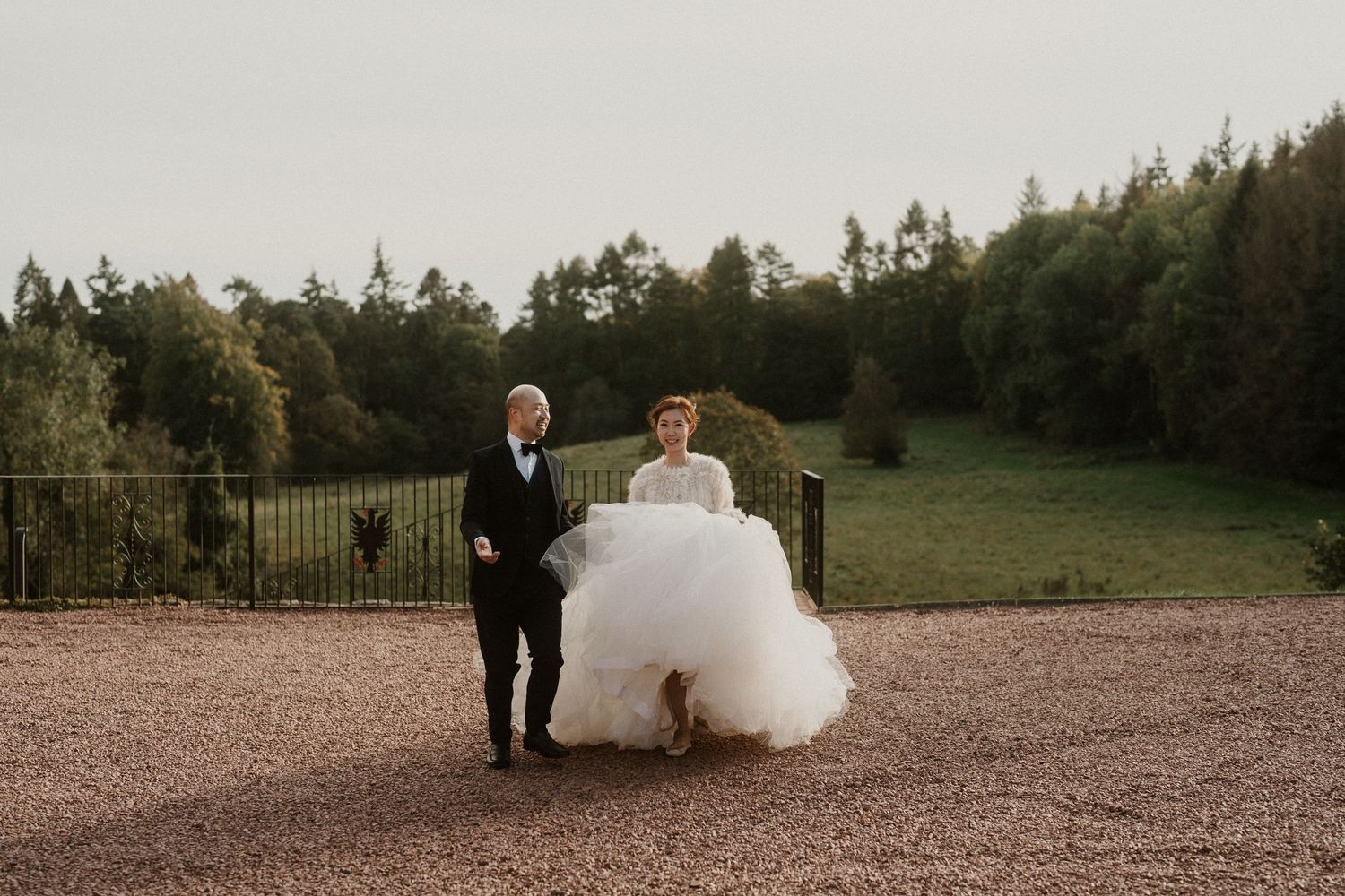 A couple embraces while dancing on a gravel path with evergreen trees in the background.