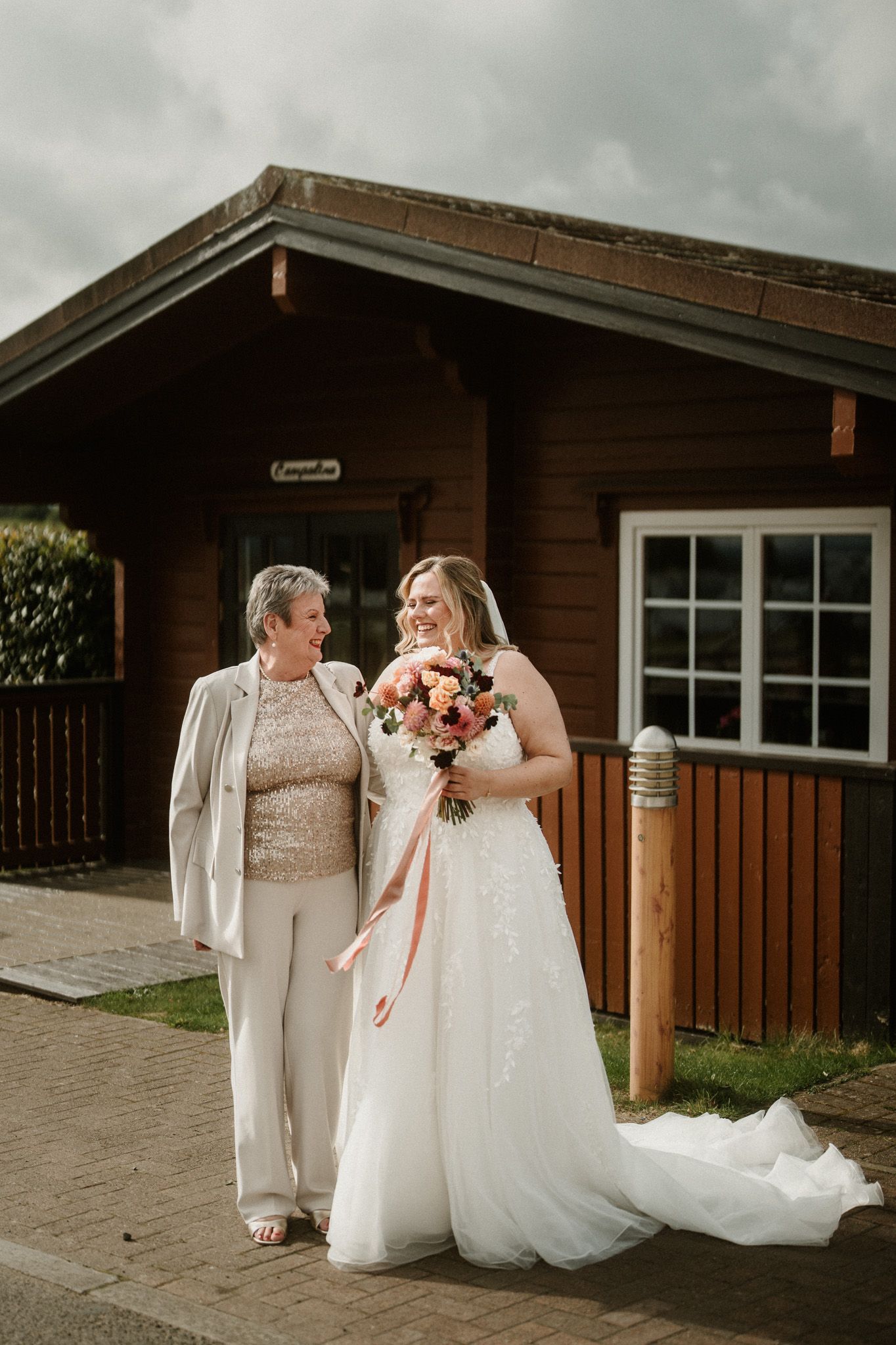 Two people in white wedding dresses share a candid moment outside a wooden lodge building.