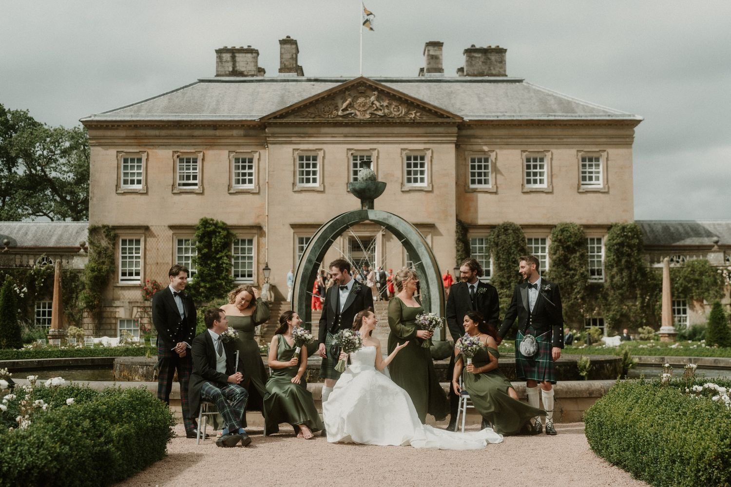 Wedding party poses in front of an elegant Georgian manor house surrounded by manicured gardens.