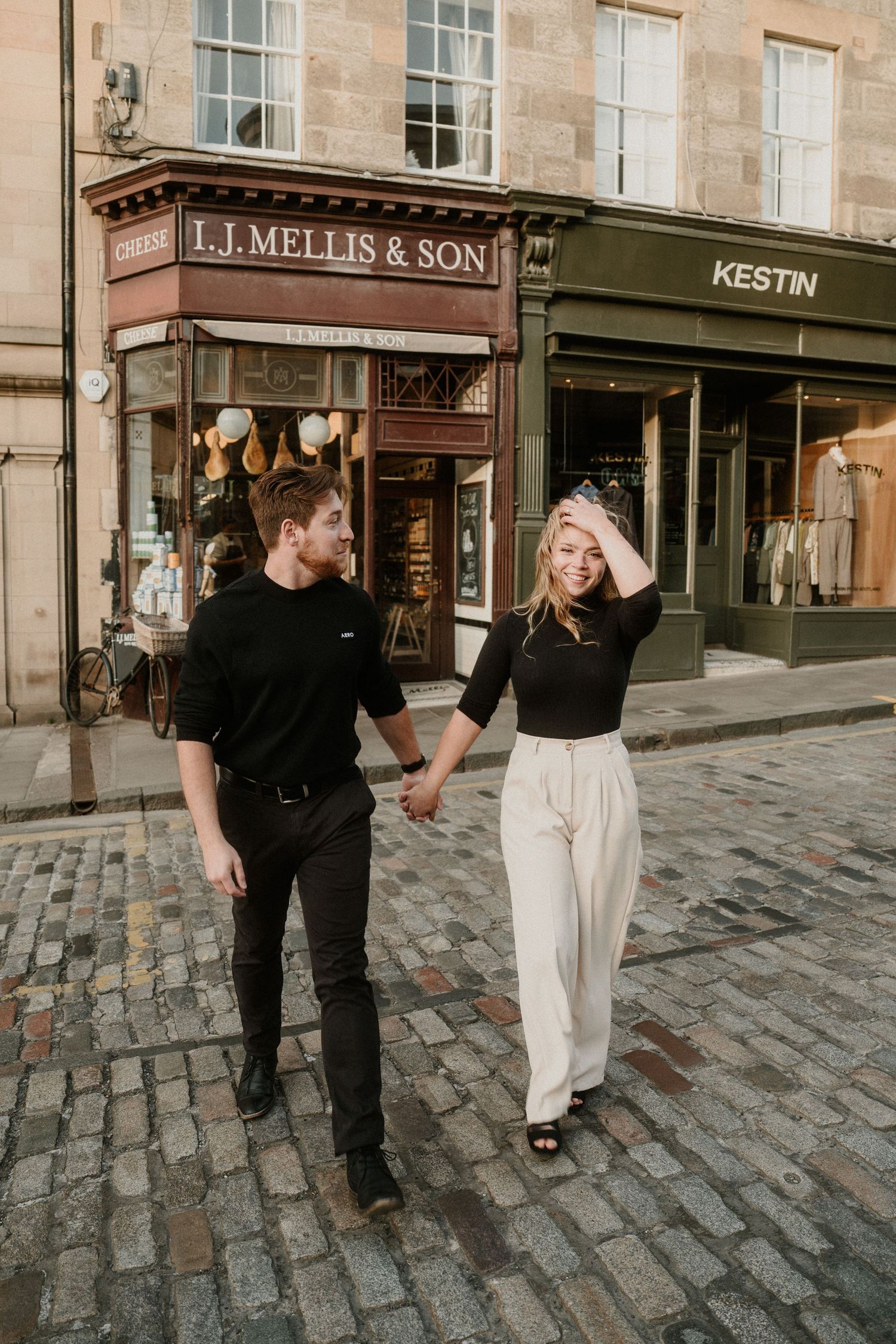 Couple holding hands walking on cobblestone street in Edinburgh past I.J. Mellis & Son and Kestin shops.