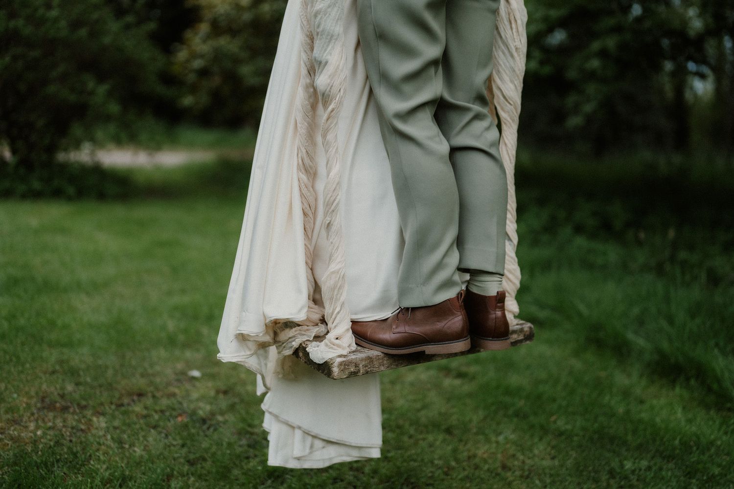 Close-up of bride in white gown and groom in sage green suit with brown leather shoes standing on lush grass.