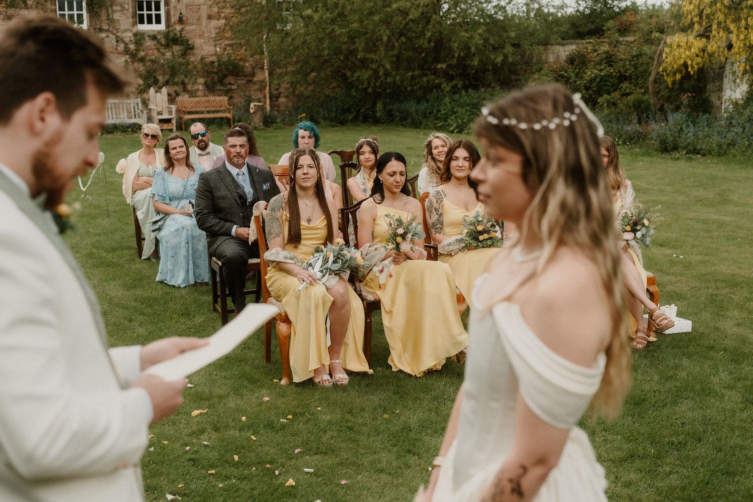 Bride and groom exchange vows outdoors with bridesmaids in yellow dresses seated on lawn at elegant garden wedding.