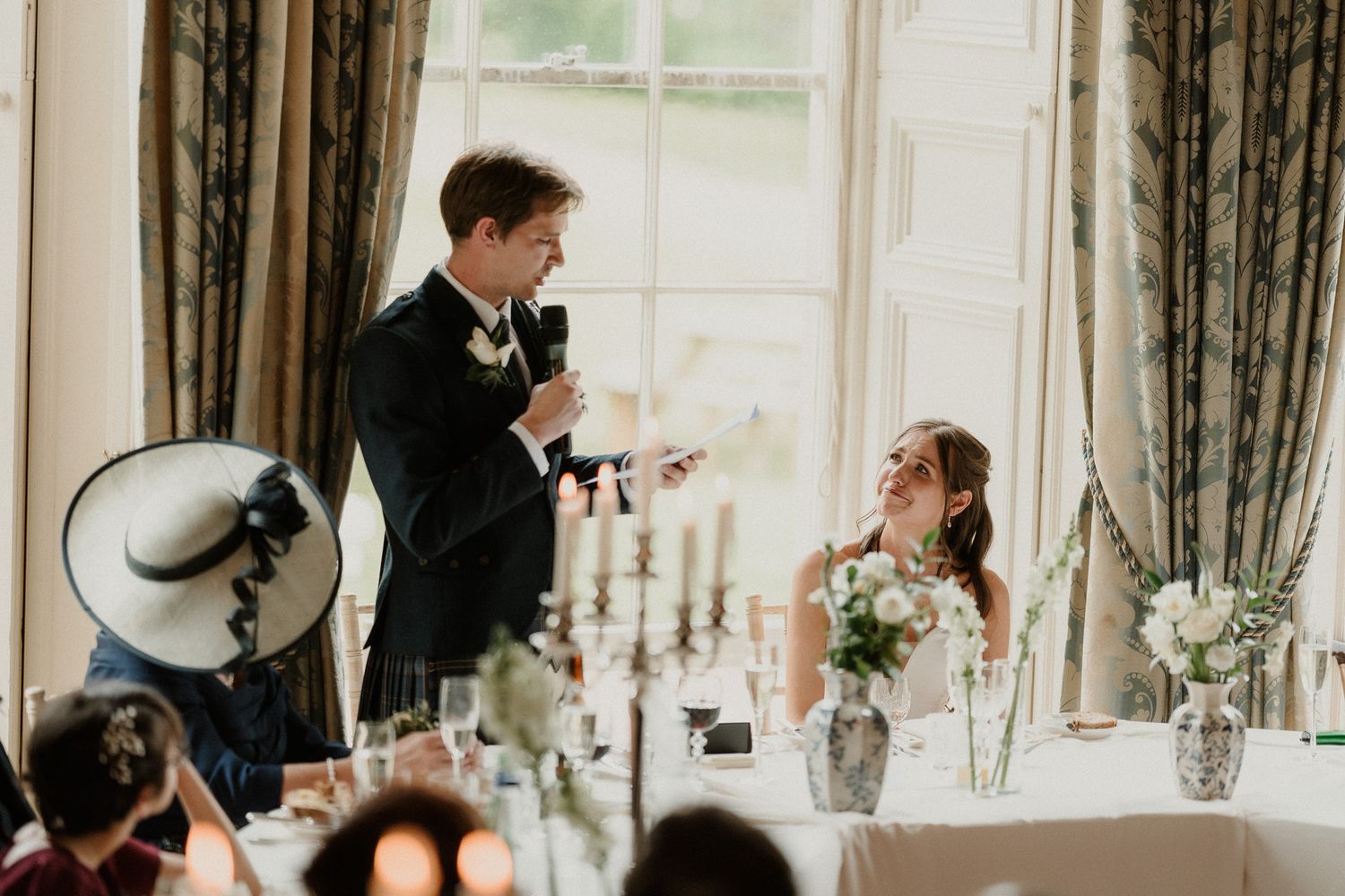 Groom giving a heartfelt wedding speech at an elegant reception, with bride smiling at the head table.