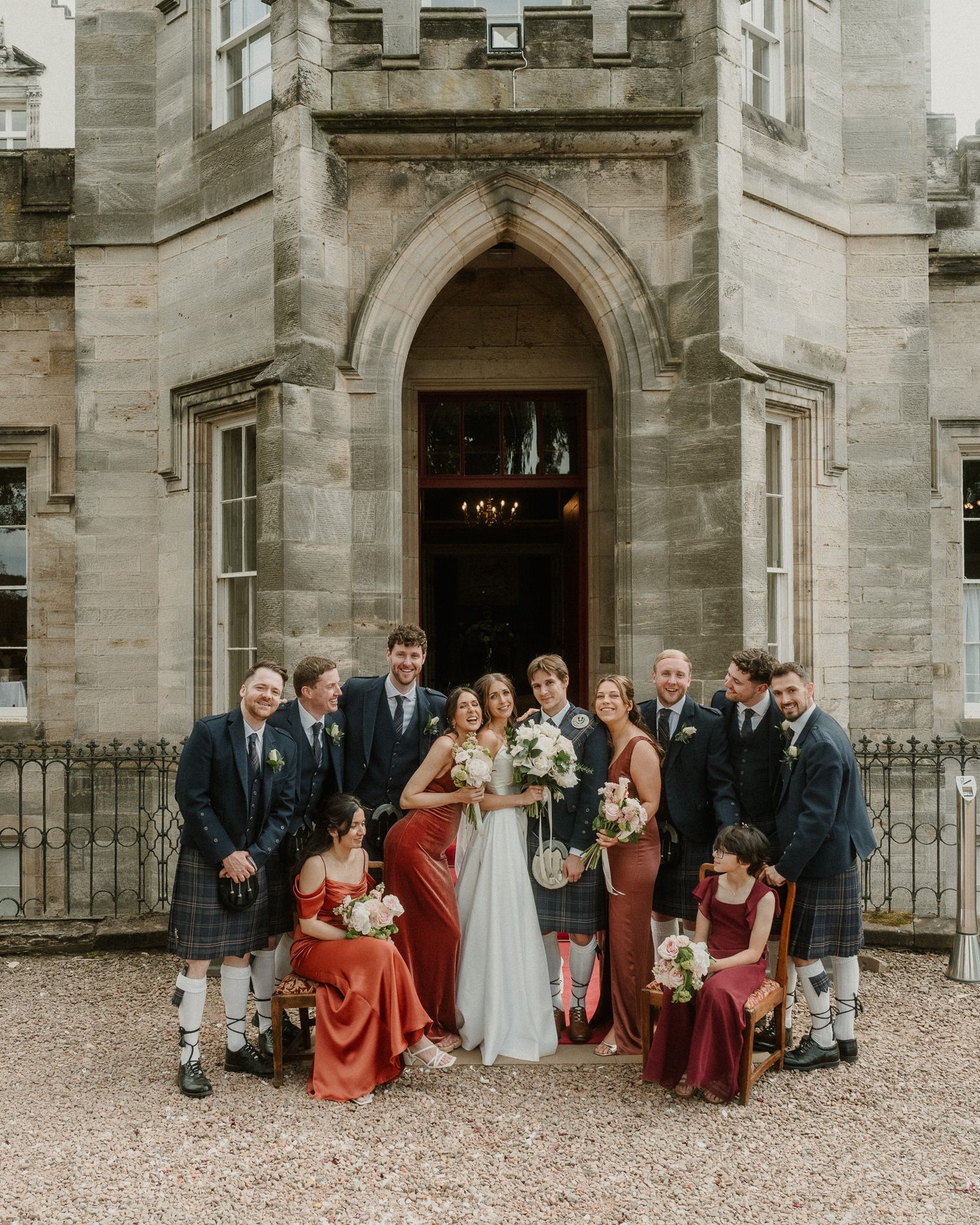 Wedding party posing in front of a Scottish castle, bride in white gown with bridesmaids in red and groomsmen in kilts.