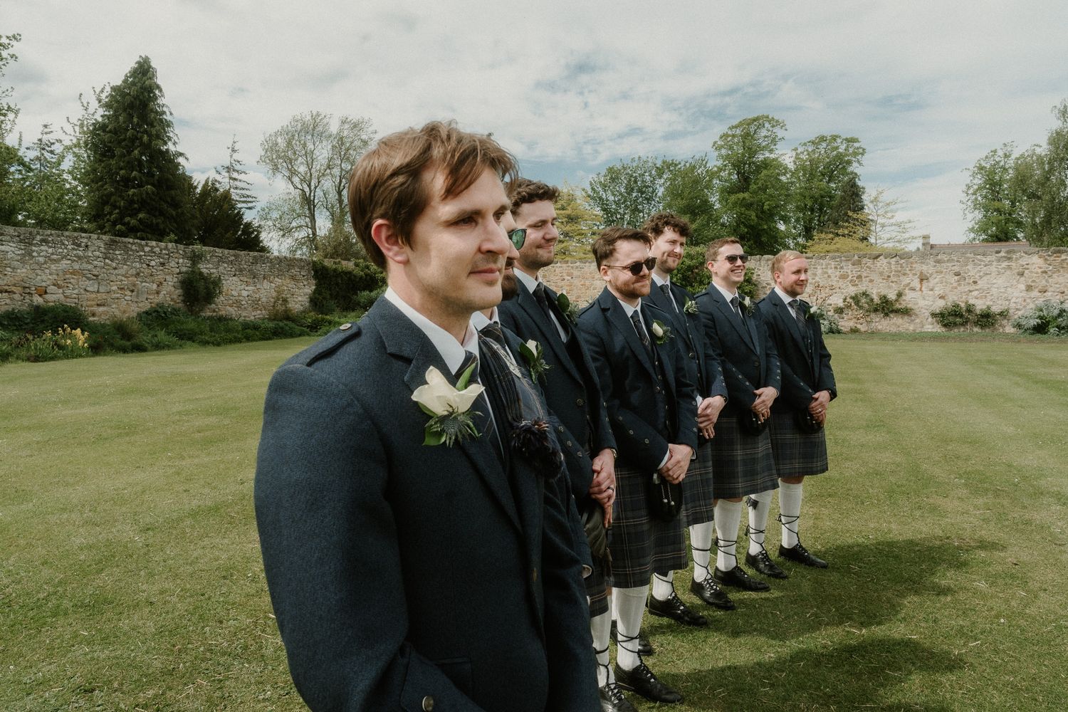 Groomsmen in traditional Scottish kilts and dark jackets standing in a row outdoors at a wedding ceremony.