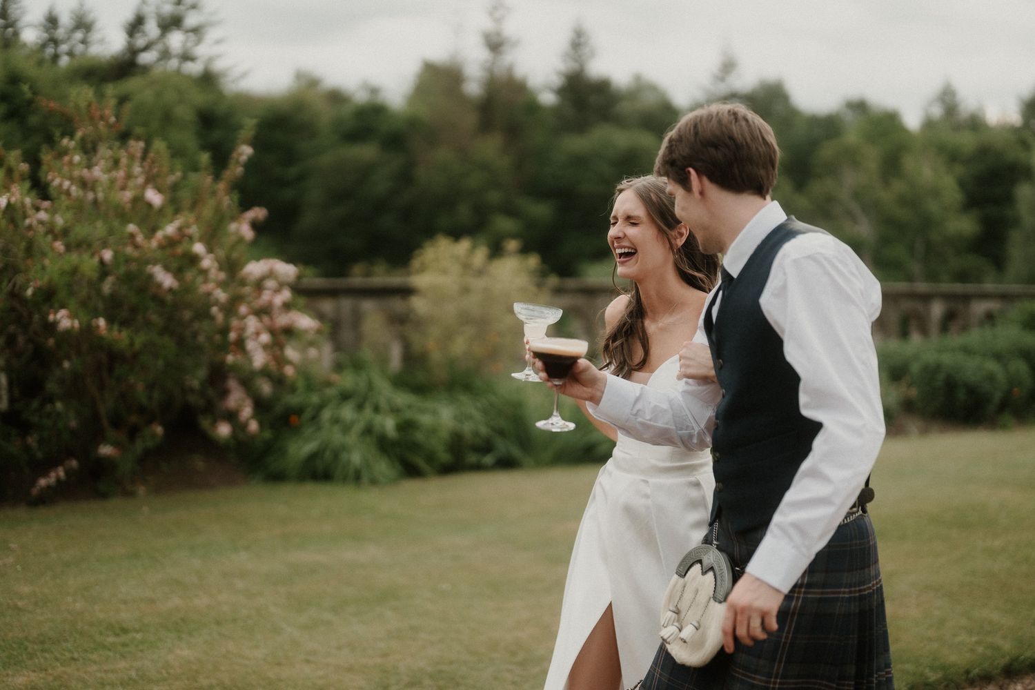 Happy bride and groom laughing outdoors, bride holding cocktail, groom in tartan kilt, lush green garden wedding setting.