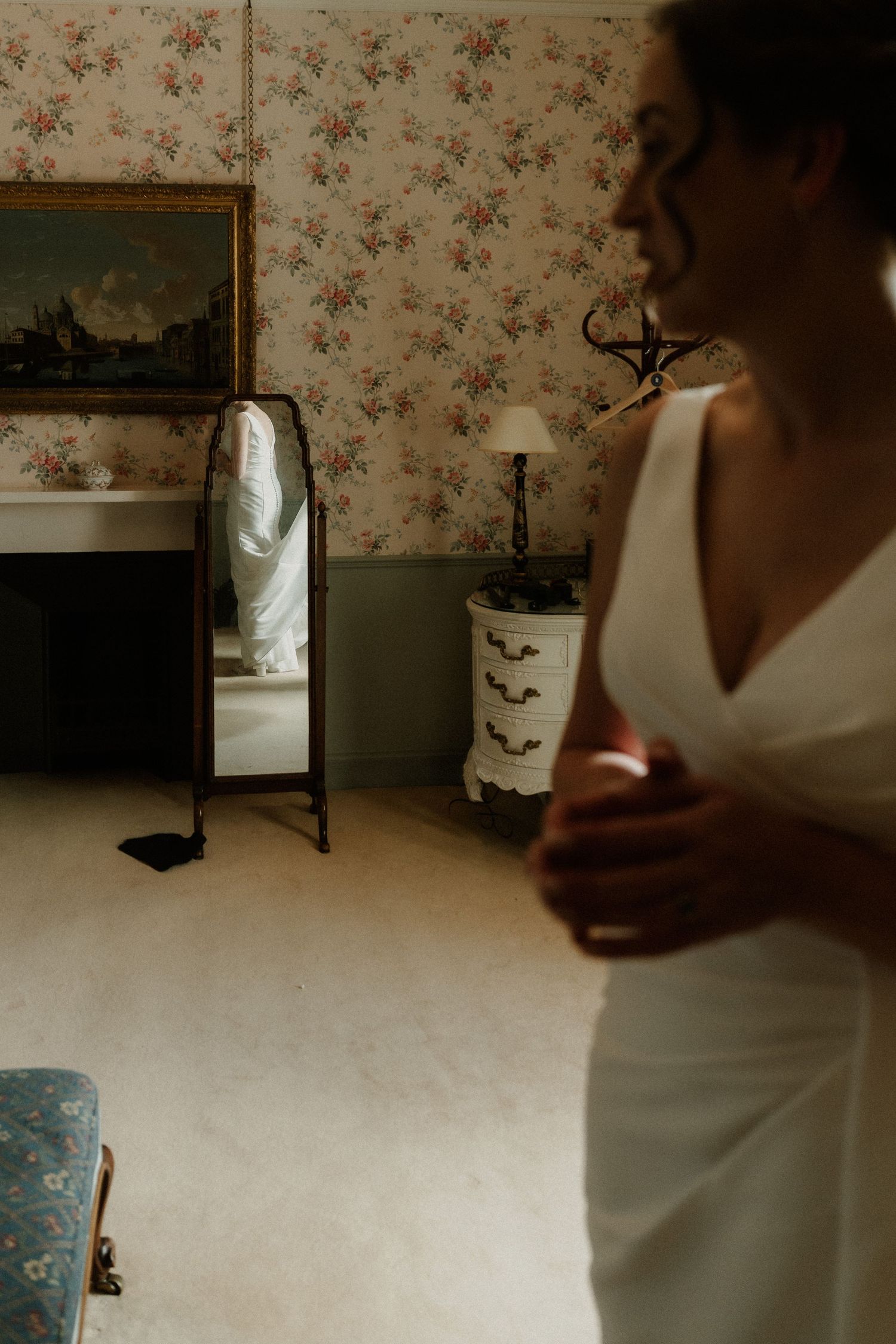 Bride in white dress stands in vintage floral wallpapered room with floor mirror reflecting her gown.