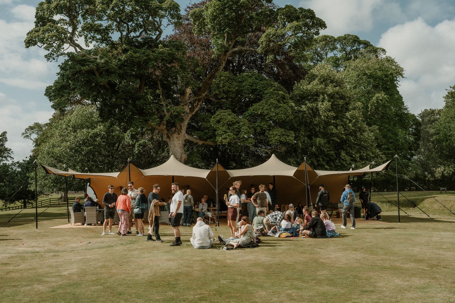 Group of people gathered under a tan stretch tent on a sunny park lawn, surrounded by large leafy trees.