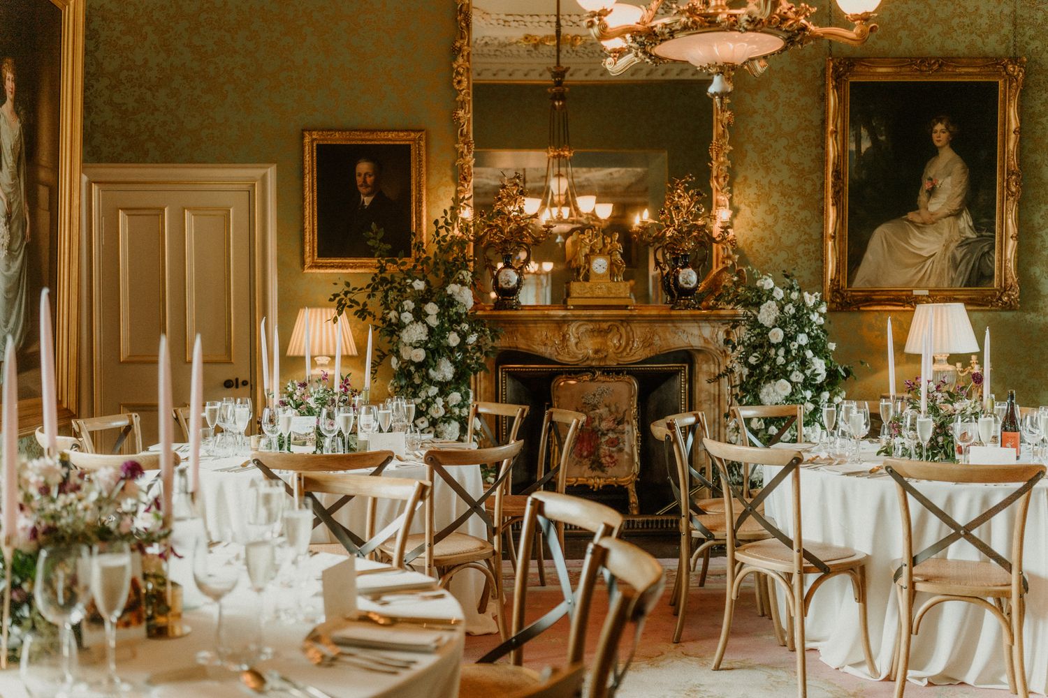 Elegant wedding reception room with candlelit tables, white floral arrangements, wooden chairs, a marble fireplace and framed portraits.