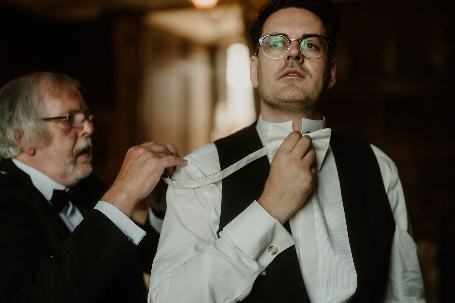 An older man in glasses helps a groom in a white shirt and black vest adjust his white bow tie before the wedding.